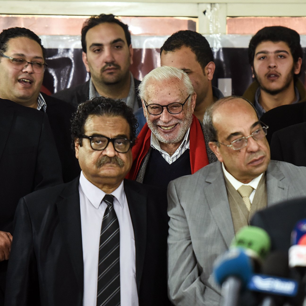 The image features a group of people gathered together, possibly at an event or press conference. There are several men in formal attire, with some wearing suits and ties, while others seem to be casually dressed. In the foreground, one man is speaking into a microphone, while others appear to be listening attentively. There is a mix of expressions among the individuals, including smiles and serious looks. The background includes a banner, suggesting this may be a significant gathering related to a specific topic or announcement.