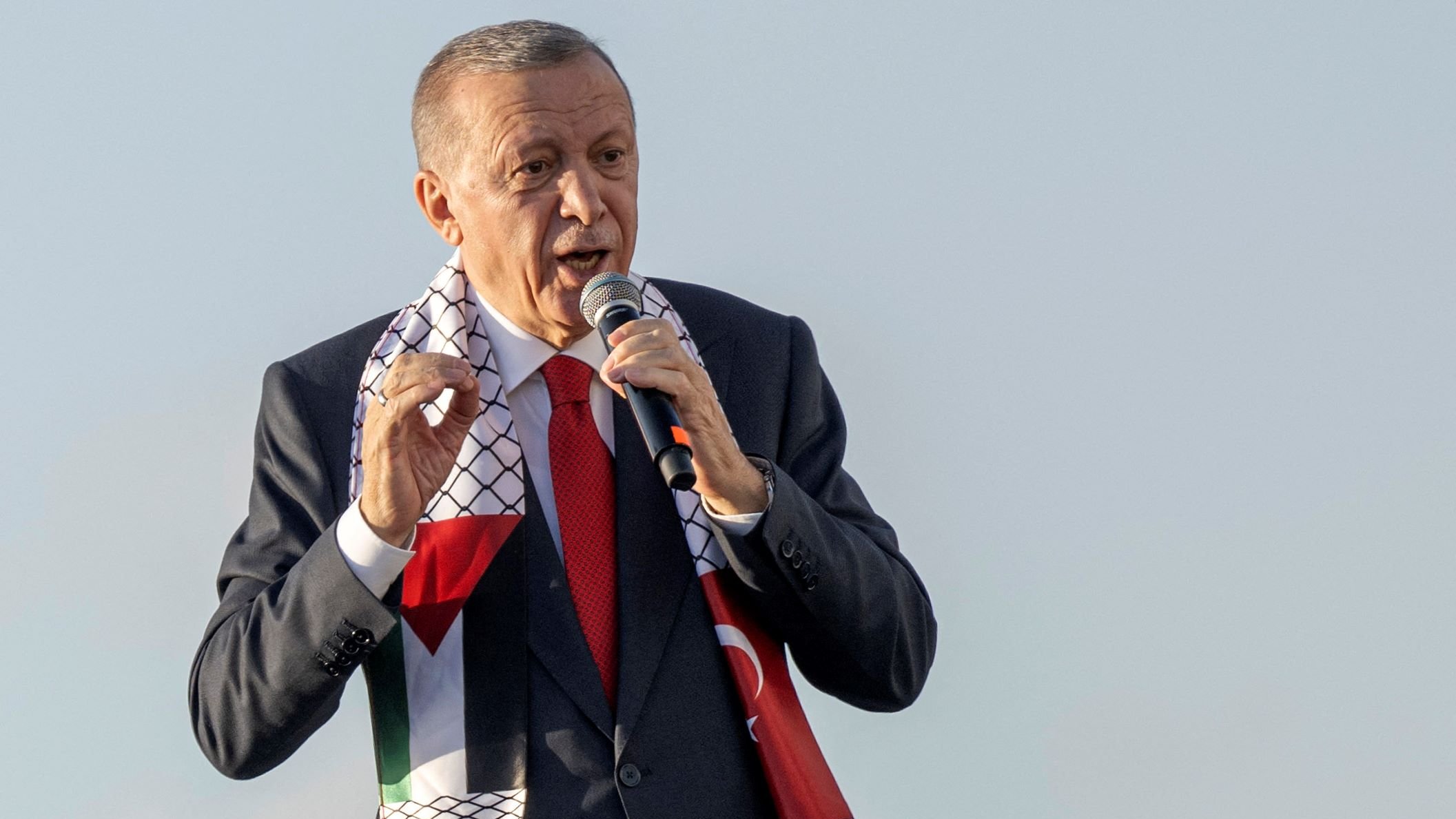 L'image montre un homme en train de parler au micro, probablement lors d'un discours ou d'une manifestation publique. Il porte un costume sombre avec une cravate rouge et un foulard aux motifs représentant la Palestine, qui est enroulé autour de son cou. L'arrière-plan semble clair, suggérant qu'il se trouve à l'extérieur. L'homme semble exprimer des émotions fortes, ce qui indique qu'il est engagé dans son discours.