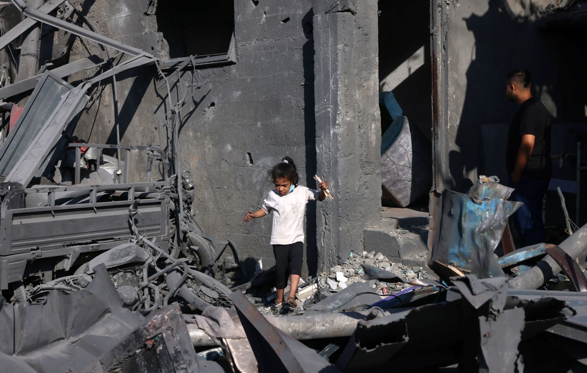 تظهر الصورة طفلاً صغيراً يرتدي الملابس البيضاء وهو يسير بين أنقاض مبنى مدمّر. الجدران حوله متهدمة ومغطاة بالرماد، مما يدل على الدمار الذي حل بالمكان. في الخلفية، يمكن رؤية شخص بالغ يقف بالقرب من الباب، يبدو وكأنه يتفحص وضع المكان. الأجواء تعكس حالة من الحزن والفوضى نتيجة الكارثة.