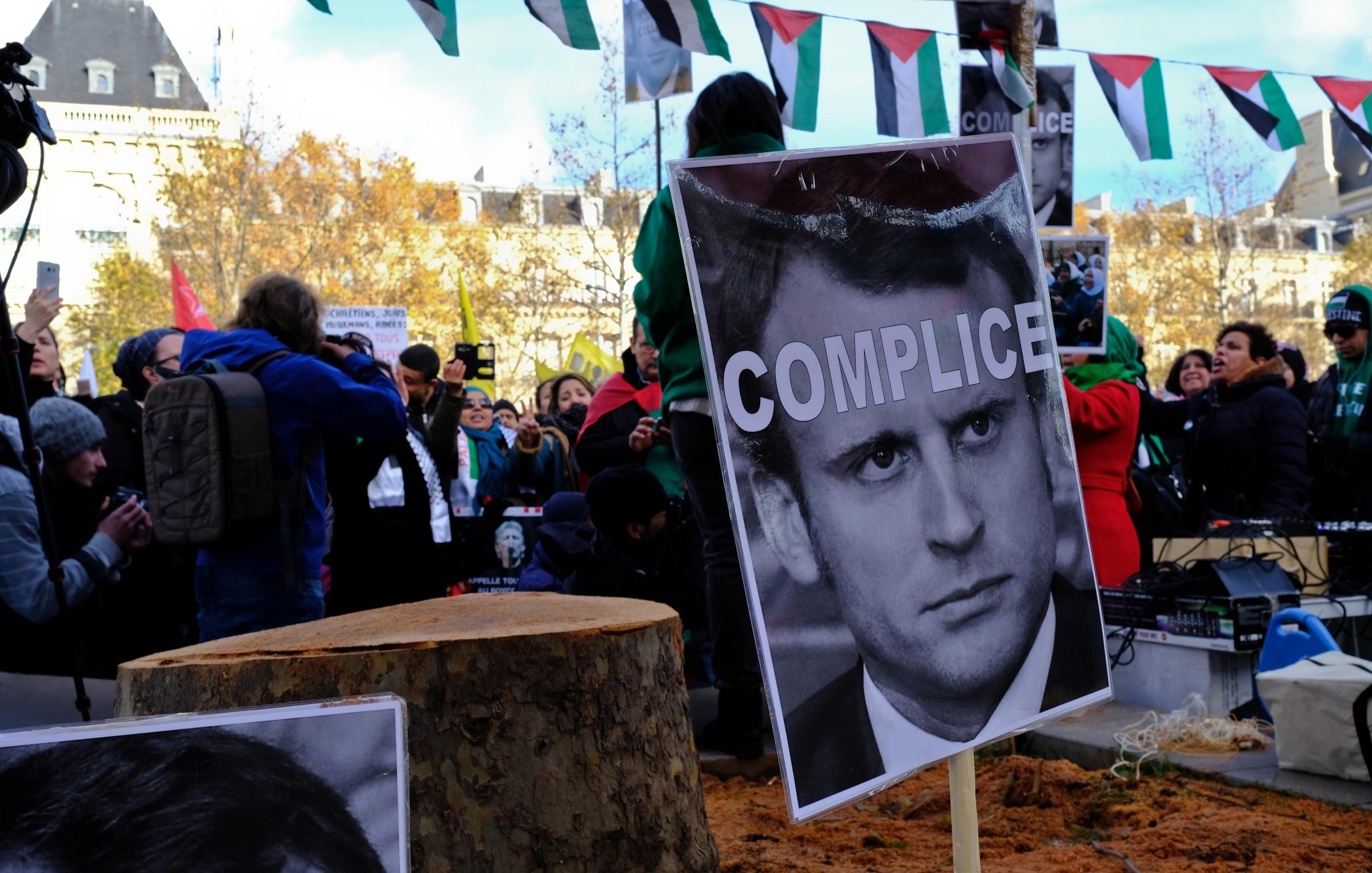 L'image montre une manifestation où une foule se rassemble autour d'un tronc d'arbre coupé. Des panneaux sont affichés avec des portraits et le mot "COMPLICE" écrit en gros caractères sur une affiche. En arrière-plan, on peut voir des drapeaux et des personnes prenant part à la protestation. L'atmosphère semble tendue, illustrant des sentiments de mécontentement et des revendications politiques.