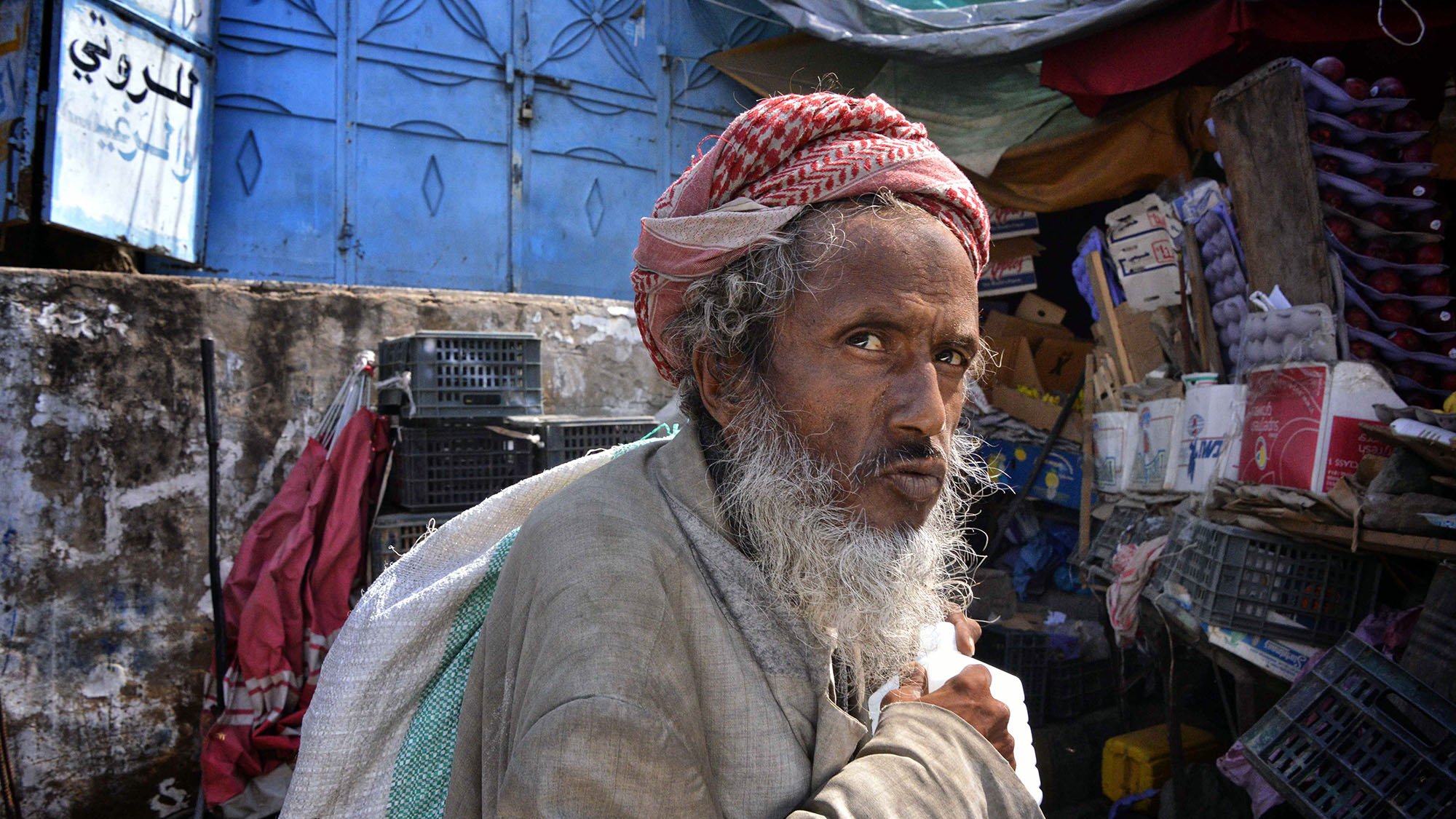 The image depicts an elderly man with a prominent beard and traditional attire, wearing a distinctive head wrap. He appears to be in a market setting surrounded by various goods and a colorful backdrop. His expression seems contemplative, and he is holding a container, possibly of milk or another liquid. The scene conveys a sense of everyday life in a bustling environment.