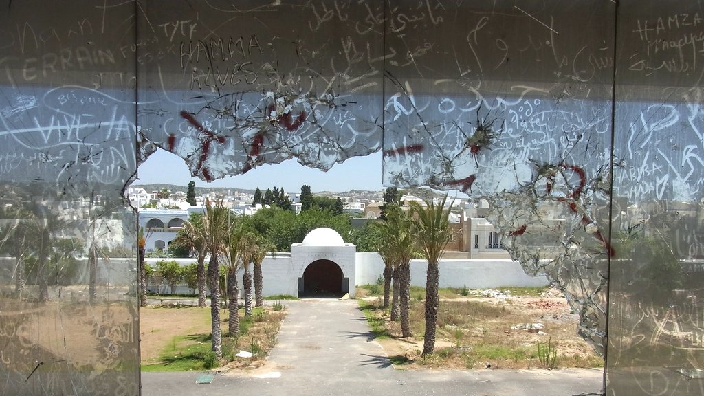 L'image montre une vue à travers un grand trou dans une vitre brisée. On peut voir un paysage urbain avec des palmiers et un édifice en arrière-plan, probablement une entrée ou un portail. Le ciel est dégagé et la scène semble être abandonnée, avec des graffiti visibles sur le verre. L'atmosphère de l'image pourrait évoquer un sentiment de désolation ou d'oubli.