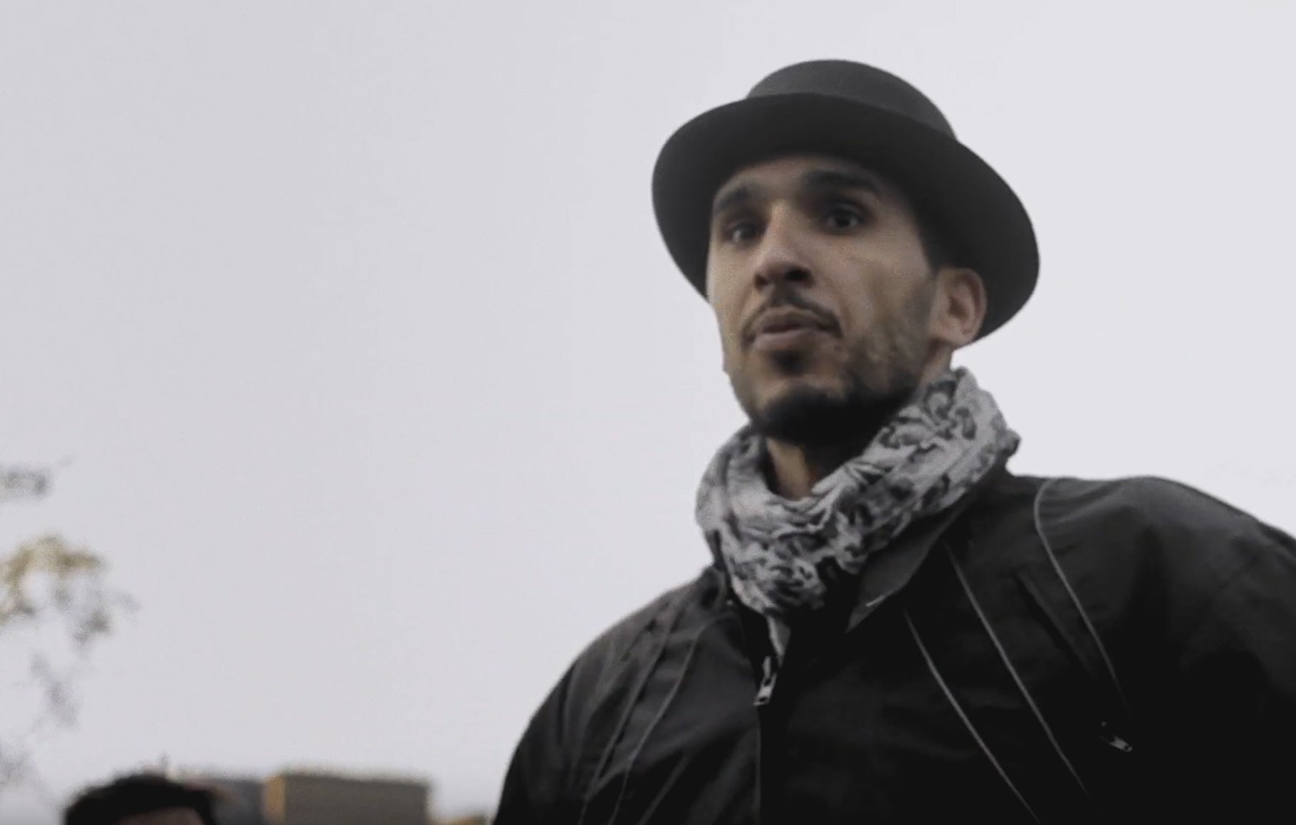 L'image montre un homme portant un chapeau noir et une écharpe. Il a une expression sérieuse et regarde droit devant lui. Le fond semble flou, suggérant une ambiance extérieure peut-être en milieu urbain ou dans un parc. Le temps semble nuageux, ce qui ajoute une atmosphère un peu sombre à la scène.