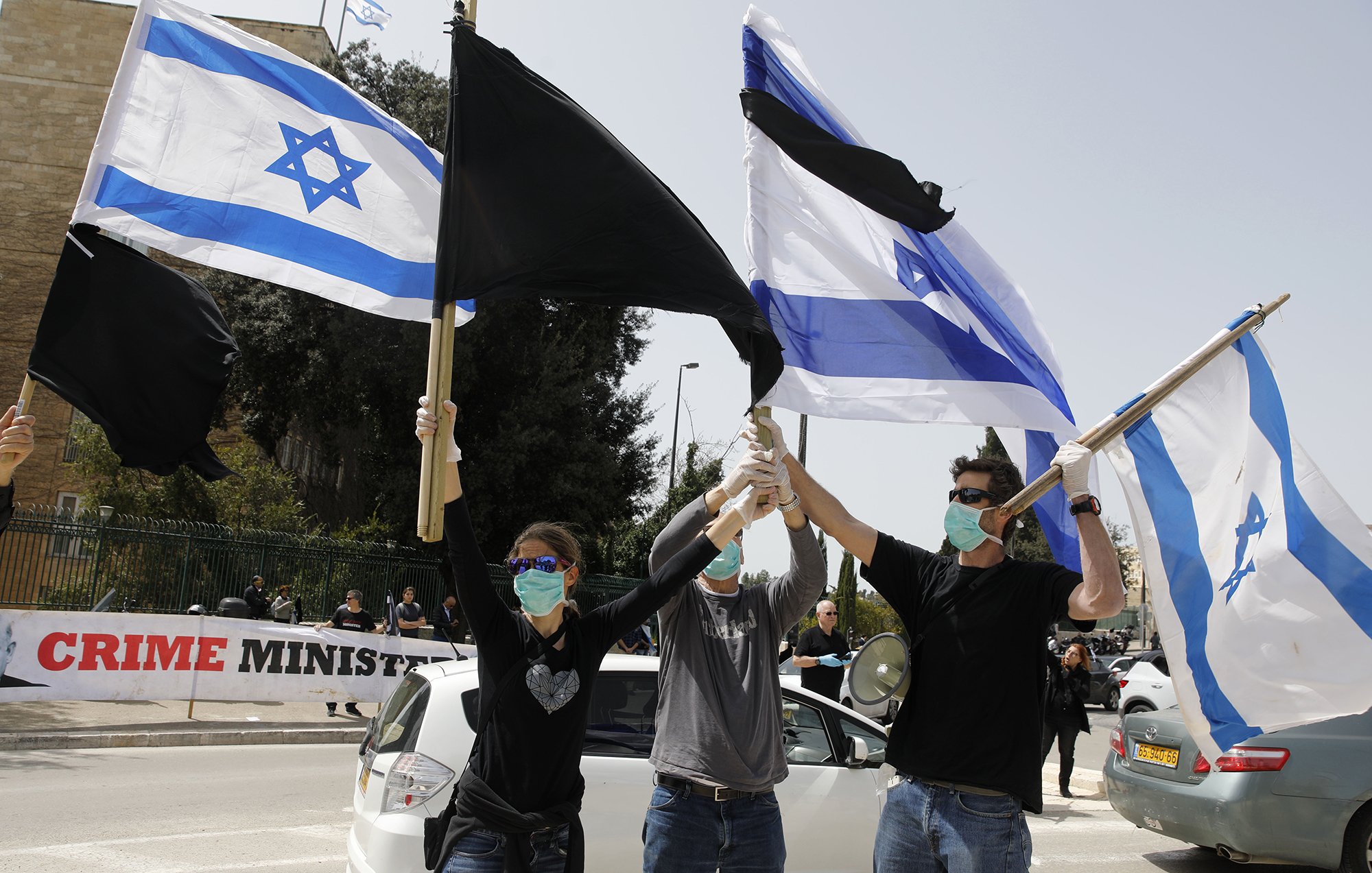L'image montre un groupe de personnes en train de manifester. Elles tiennent des drapeaux, y compris le drapeau israélien et un autre drapeau aux couleurs noire et blanche. Tous les participants portent des masques sur le visage, ce qui suggère qu'ils prennent des précautions sanitaires. En arrière-plan, on peut voir une banderole qui semble contenir un message critique envers le gouvernement. L'atmosphère semble être celle d'une protestation pacifique.