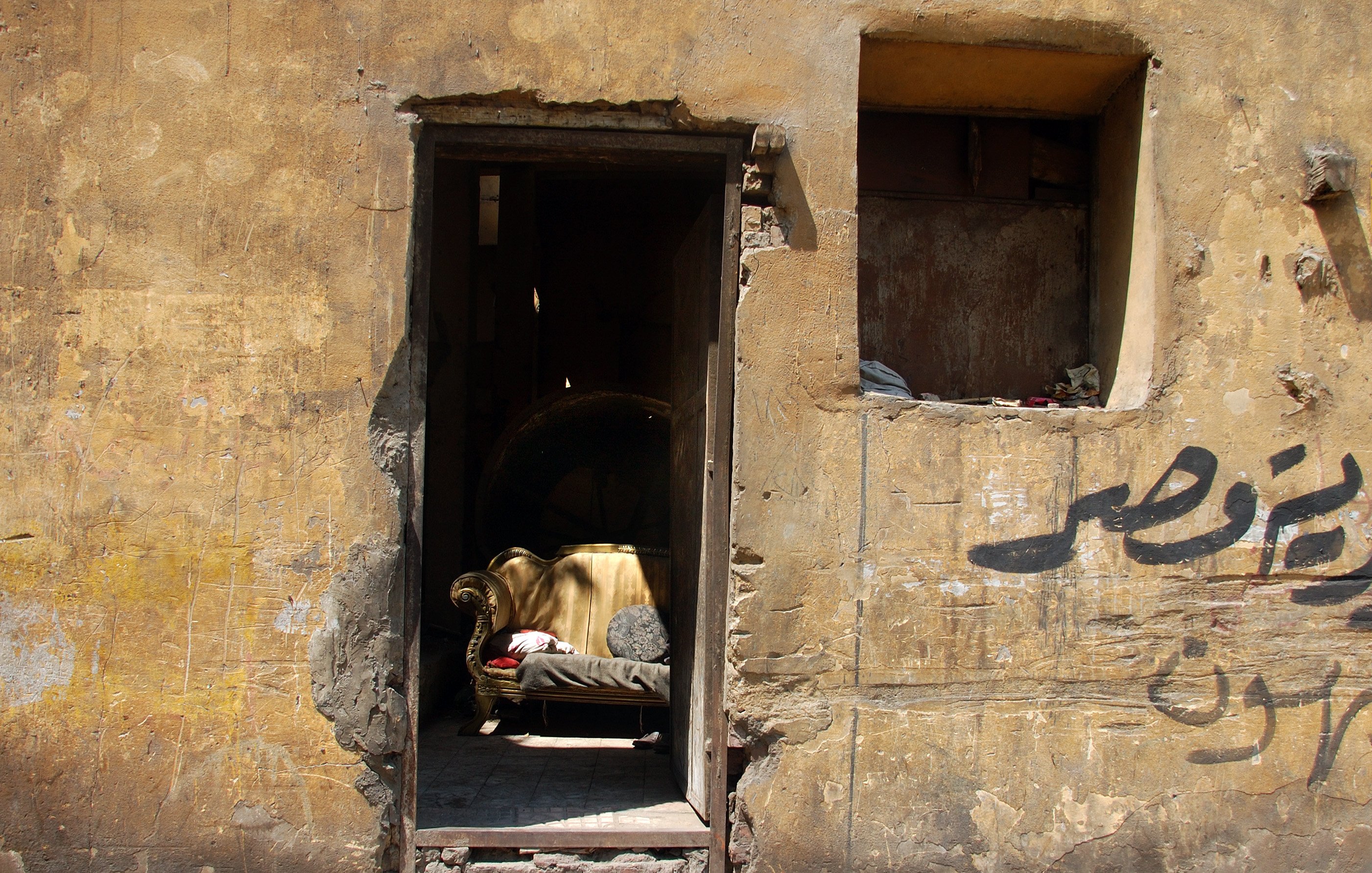 تصویر نمایی از یک درب ورودی به یک ساختمان قدیمی است که دیوارهای آن رنگ و بافت زرد و کهنه‌ای دارند. در وسط در، یک کاناپه قدیمی و ساده قرار دارد که به نظر می‌رسد سال‌ها بدون استفاده مانده است. در سمت چپ تصویر، بخشی از دیوار به صورت تخریب شده دیده می‌شود و یک پنجره کوچک بالای درب نیز وجود دارد که نیمی از آن به طور کامل تخریب شده است. روی دیوار نوشته‌هایی به زبان عربی دیده می‌شود که پس‌زمینه‌ای از فضای شهری و قدیم را به تصویر می‌کشد. کل صحنه حس نوستالژیک و متروکه‌ای را منتقل می‌کند.