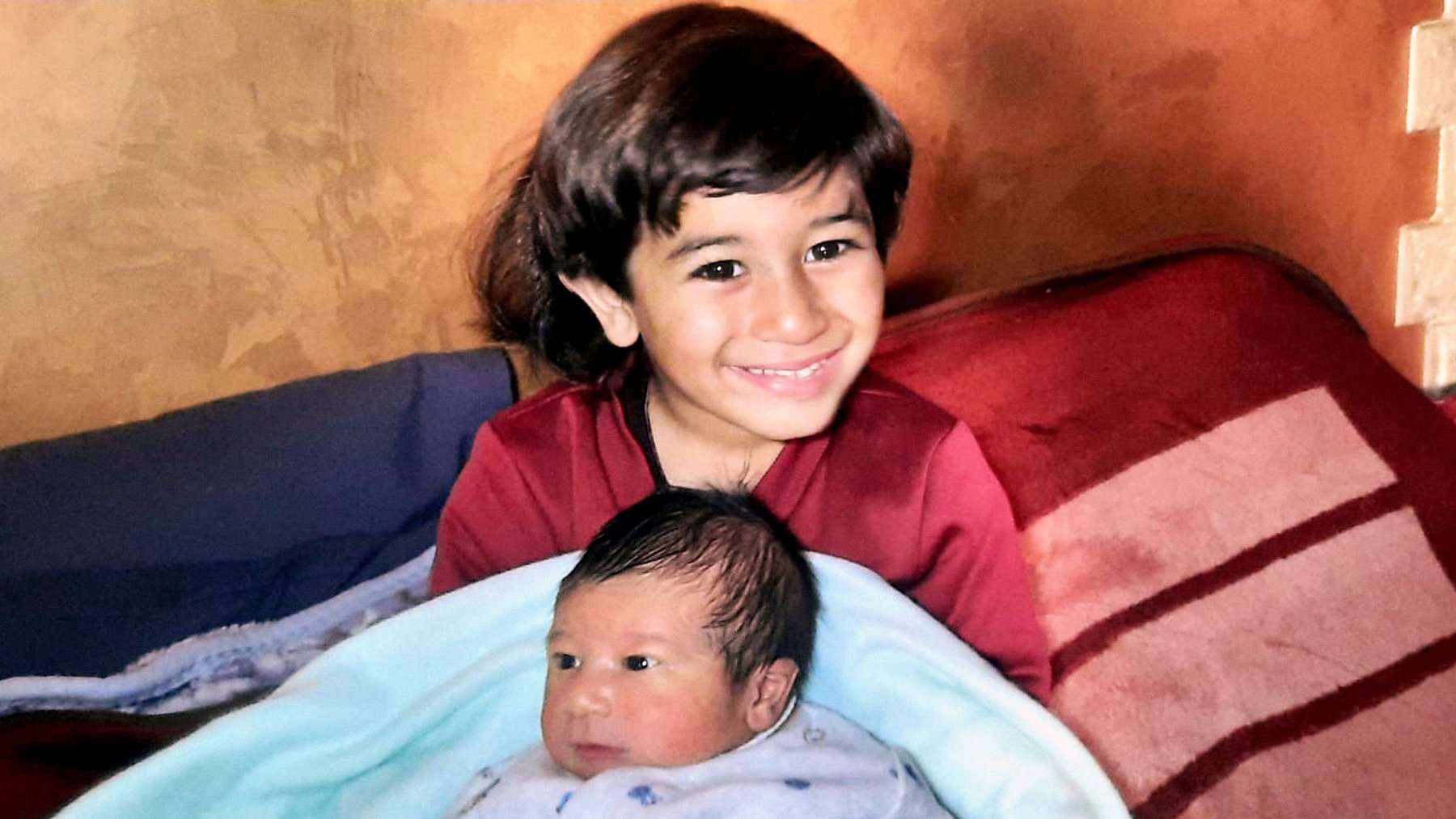 Nell'immagine si vede un bambino più grande che sorride mentre tiene in braccio un neonato avvolto in una coperta blu. Il bambino più grande ha i capelli scuri e lunghi, e indossa una maglietta rossa. Il neonato ha i capelli scuri e un'espressione curiosa. Sullo sfondo ci sono delle coperte sul lettino, creando un'atmosfera accogliente e familiare.