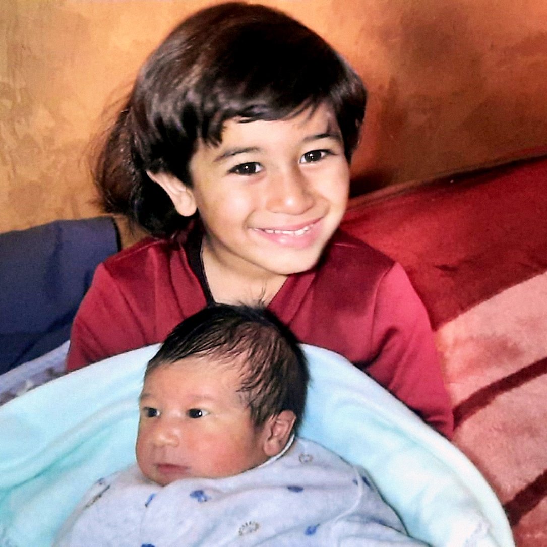 L'image montre un enfant souriant assis sur un canapé, tenant un bébé enveloppé dans une couverture bleue. L'enfant plus âgé semble heureux et fier, tandis que le bébé a une expression calme. Le décor est chaleureux avec des murs aux teintes terreuses.