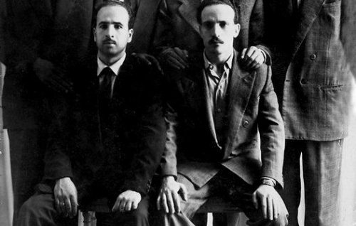 L'image montre un groupe de six hommes, habillés en costumes, posant ensemble. Ils sont disposés sur deux niveaux : trois debout à l'arrière et trois assis à l'avant. Tous les hommes affichent une expression sérieuse et se tiennent près les uns des autres, ce qui donne une impression d'unité. L'arrière-plan est neutre, avec des rideaux qui ajoutent une texture à la scène. La photo semble être en noir et blanc, ce qui lui confère un aspect vintage.