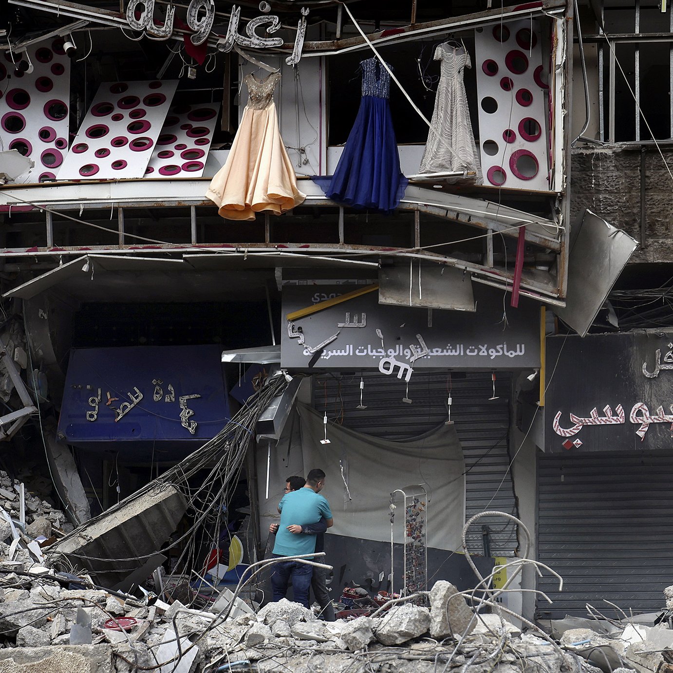 L'image montre des débris d'un bâtiment endommagé, probablement en raison d'un conflit ou d'une catastrophe. À l'avant, on peut voir des robes suspendues à l'extérieur d'une boutique, tandis qu'en bas, deux personnes semblent fouiller ou inspecter les lieux. Les murs sont en ruine, et l'environnement dégage une impression de destruction et de désolation. Des enseignes de magasins sont visibles, mais partiellement masquées par les débris.