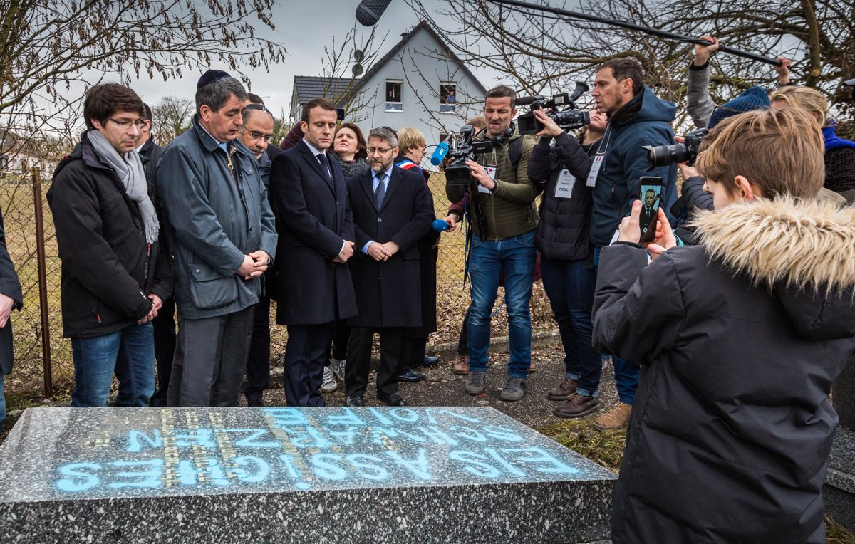 L'image montre un groupe de personnes rassemblées autour d'une stèle ou d'une tombe. Parmi elles, on peut distinguer des individus en tenue formelle, semblant assister à une cérémonie hommage. Un enfant tient un téléphone pour filmer ou prendre une photo. Des journalistes, avec des caméras et des micros, sont également présents, suggérant que l'événement est médiatisé. En arrière-plan, on aperçoit une maison et des arbres, indiquant un cadre extérieur. 