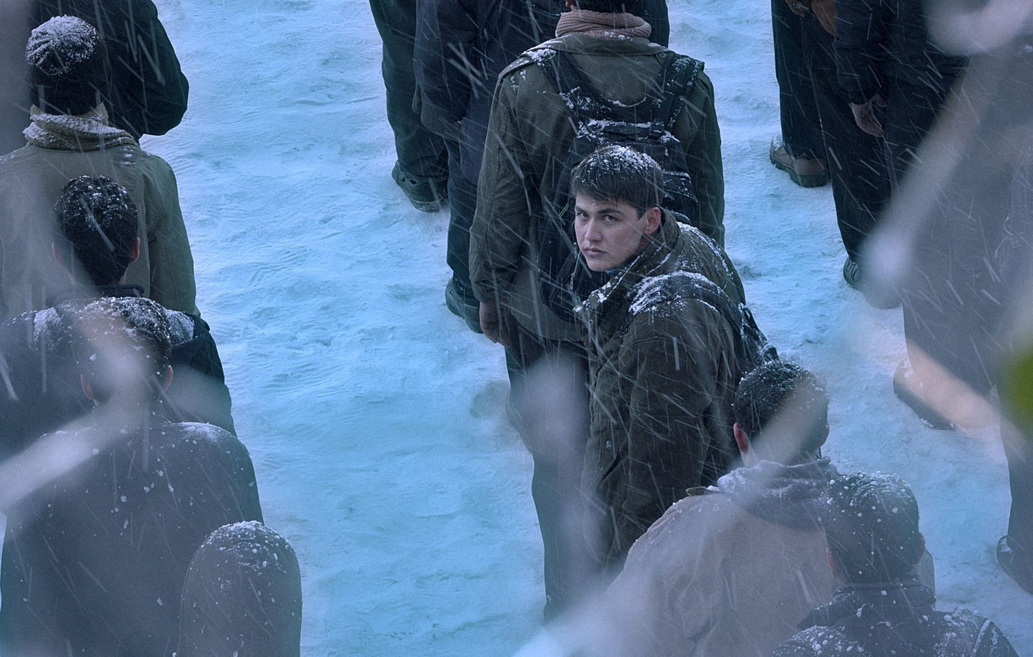 L'image montre une scène froide et enneigée avec une foule de personnes, des flocons de neige tombant. Au centre, un jeune homme se retourne, son visage est légèrement visible. Les autres autour de lui sont flous et vêtus de manteaux sombres, créant une atmosphère de tension et d'incertitude.