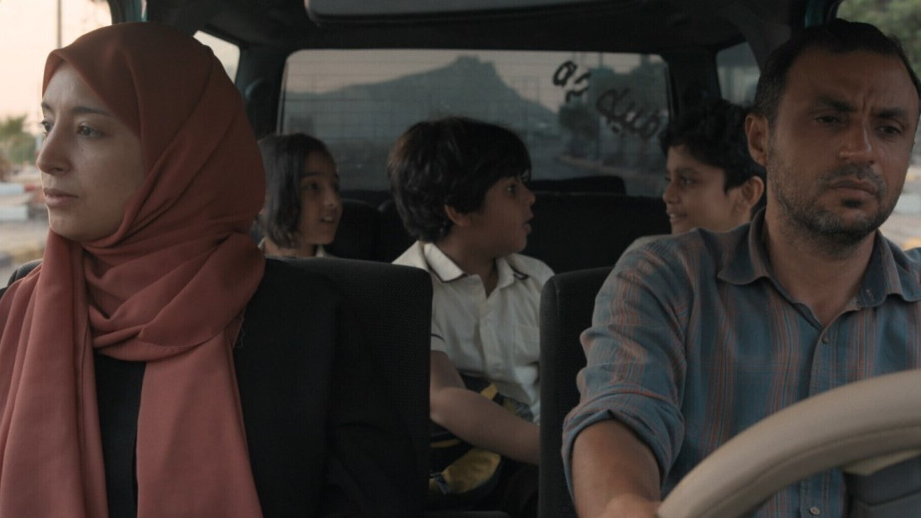 L'image montre un groupe de personnes dans une voiture. À l'avant, un homme au visage sérieux conduit, tandis qu'une femme portant un hijab est assise à côté de lui, semblant pensive. À l'arrière, deux enfants et un adolescent se parlent, créant une atmosphère de dynamisme et d'interaction. La lumière semble douce, suggérant une scène de fin de journée. L'ambiance générale évoque une sorte de voyage ou de déplacement, avec un mélange d'émotions au sein de la voiture.