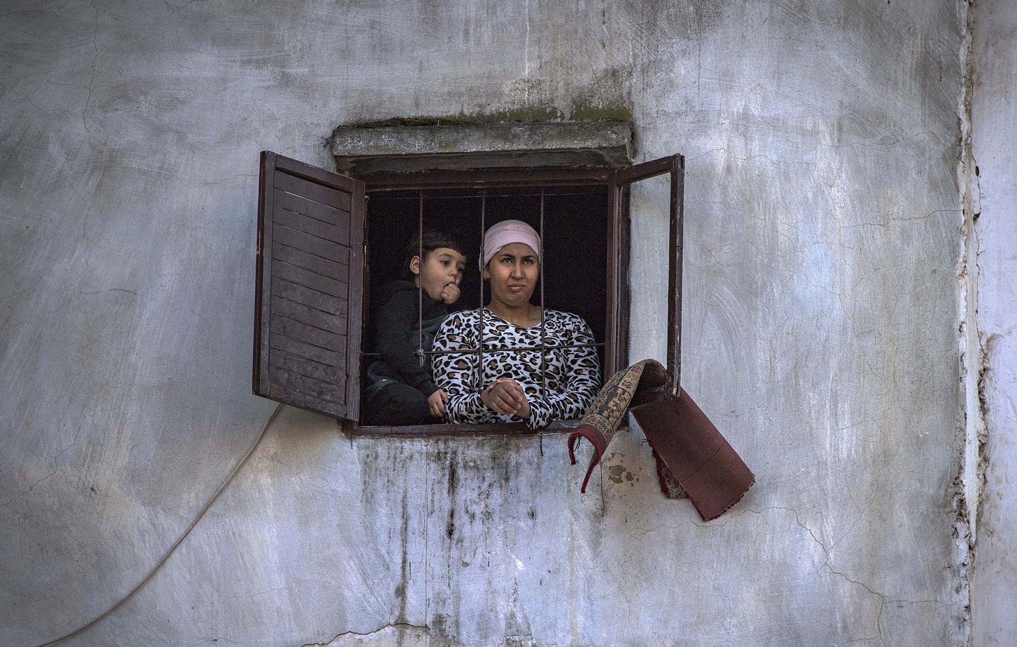 L'image montre deux personnes regardant à travers une fenêtre. Il y a une femme, vêtue d'un haut à motifs, qui semble attentive. À côté d'elle, un enfant, visiblement curieux, se penche légèrement. L'environnement est simple, avec des murs en crépi et une fenêtre aux volets ouverts. L'atmosphère semble réfléchie et calme.