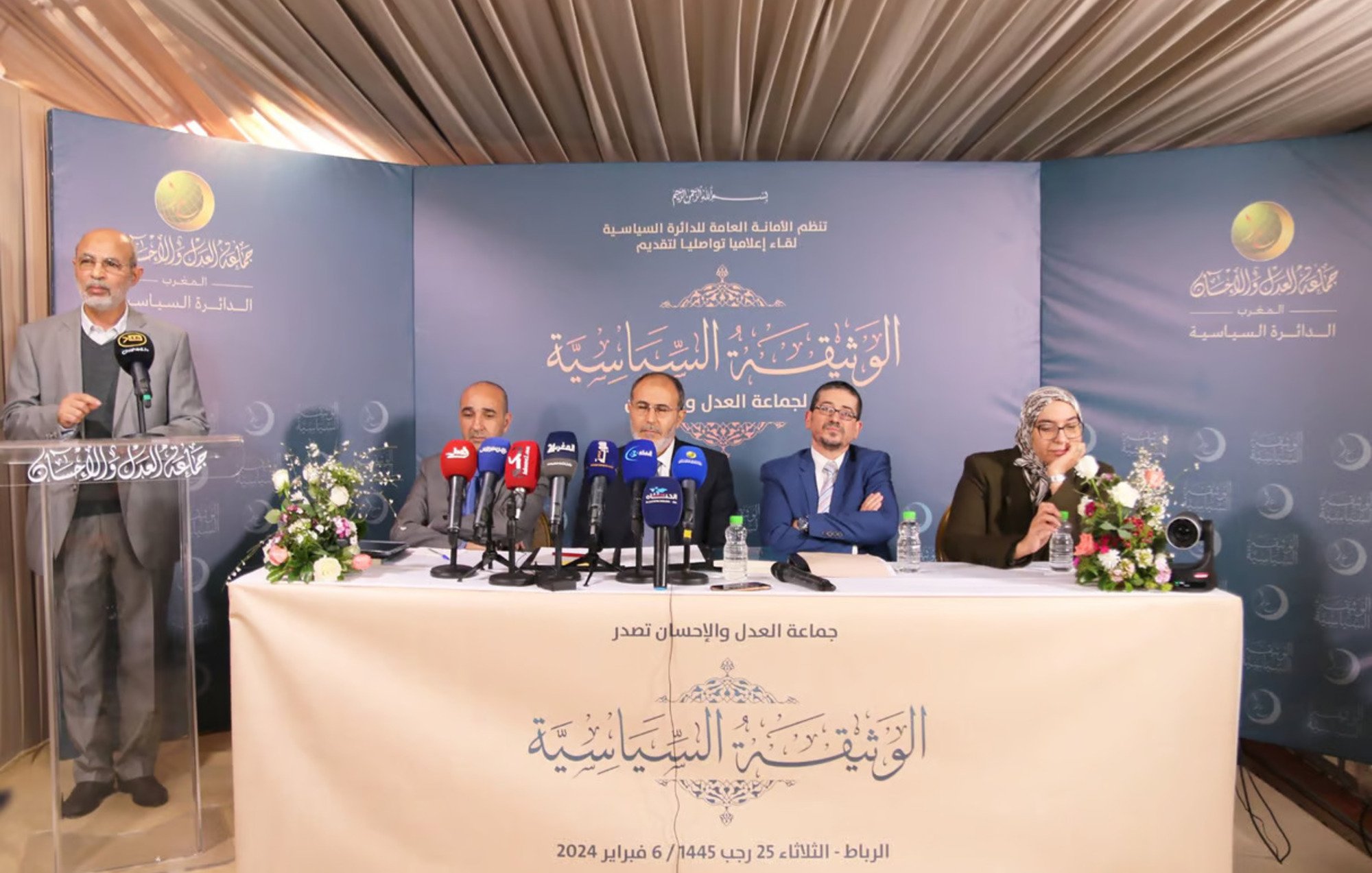 الصورة تظهر مؤتمرًا صحفيًا أو اجتماعًا رسميًا. يوجد على المنصة أربعة أشخاص يجلسون معًا، حيث يتحدث أحدهم ويبدو أنه يتحدث في ميكروفون. هناك لافتة خلفهم تحمل عنوان الاجتماع، ومجموعة من الكاميرات والشاشات تسجل أو تنقل الحدث. الطاولة مزينة بالزهور، والإضاءة هادئة. الخلفية تبدو أنيقة وتعكس الجدية الرسمية للاجتماع.