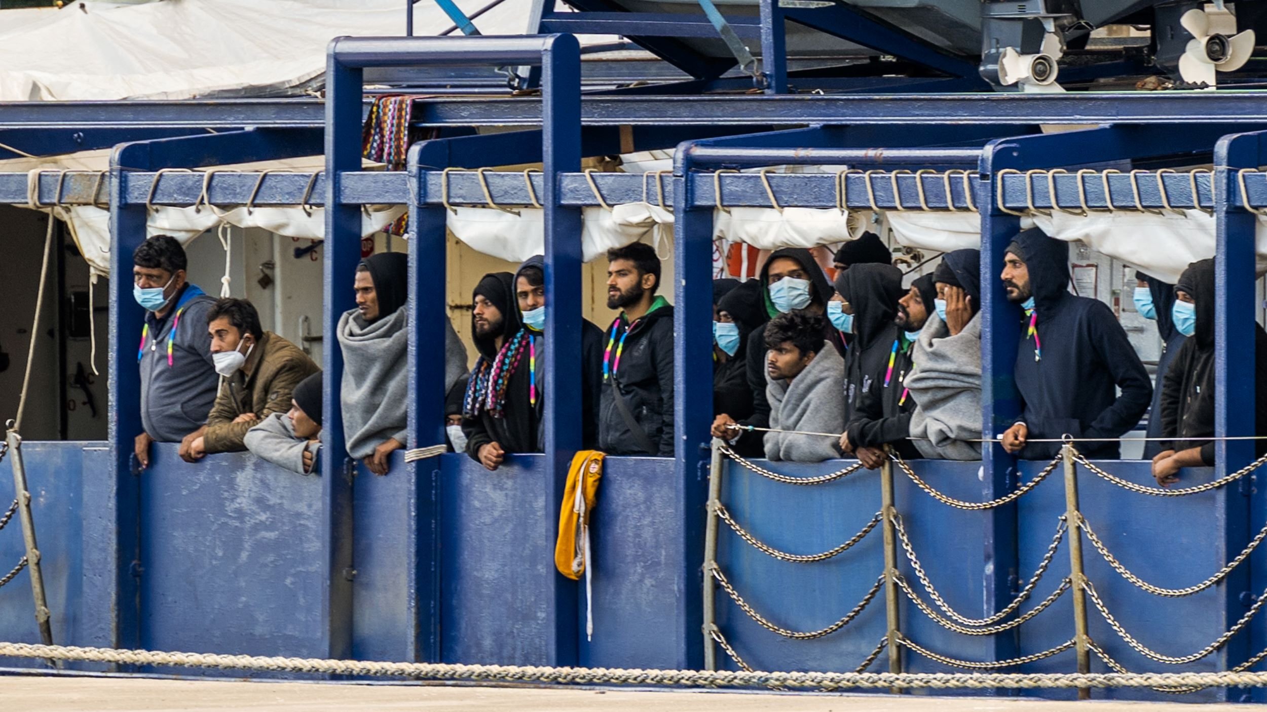 تظهر الصورة مجموعة من الأشخاص يقفون على متن عبارة أو سفينة. يرتدي بعضهم أغطية رأس، بينما يرتدي الآخرون معاطف أو ملابس دافئة. يبدو أن الجميع يشاهدون شيئًا ما في الأفق، ويبدو عليهم القلق أو الترقب. كما أنهم يرتدون كمامات، مما يشير إلى أنهم قد يكونوا في منطقة تتطلب اتخاذ احتياطات صحية. الخلفية تحتوي على أجزاء من السفينة وأجزاء من الرصيف.