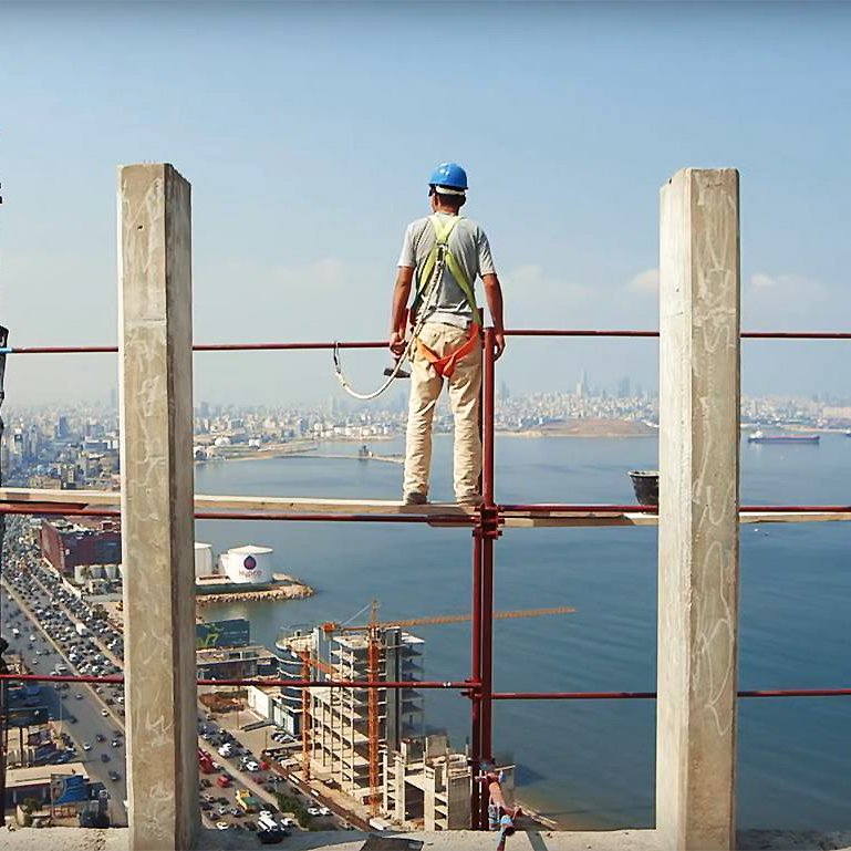 L'image montre un ouvrier de la construction se tenant sur un échafaudage, surplombant une vue panoramique sur une ville côtière. Il porte un casque de sécurité et des vêtements de travail. À l'arrière-plan, on peut voir des immeubles, une route qui longe la mer, ainsi que des bateaux sur l'eau. Le ciel est dégagé, laissant entrevoir une belle journée. La scène évoque à la fois le travail ardu des ouvriers et une magnifique perspective sur l'urbanisme et la nature environnante.