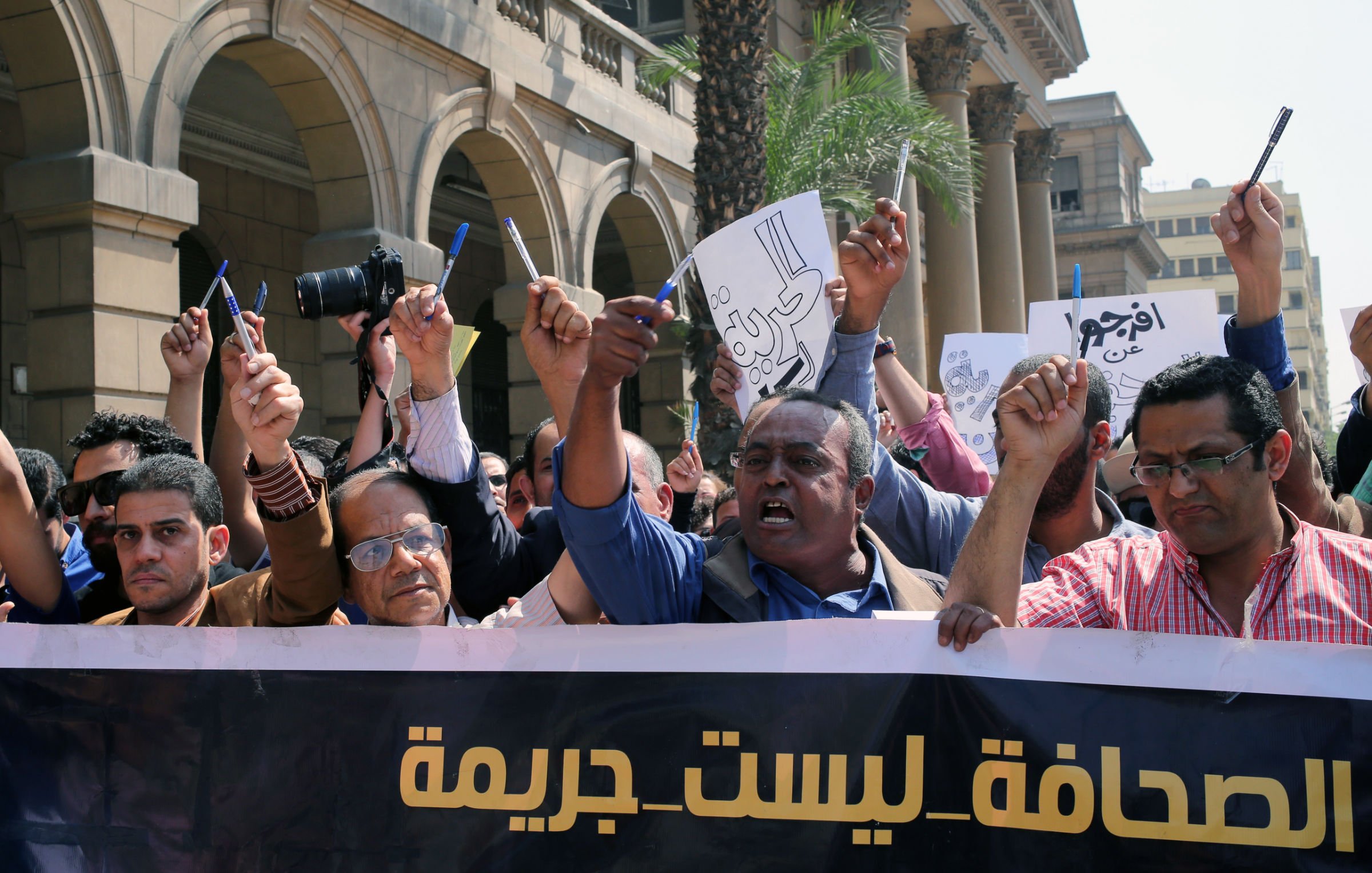 تظهر في الصورة مظاهرة تضم مجموعة من الأشخاص يحملون لافتات وأعلامًا. يرفع البعض أيديهم بشكل تعبيري، بينما يبدو البعض الآخر يتحدث أو يحتج. يوجد شعارات على اللافتات تعبر عن قضايا تتعلق بالصحافة وحرية التعبير، حيث يظهر نص مكتوب يشير إلى أن الصحافة ليست جريمة. الخلفية تحتوي على مباني كبيرة، مما يشير إلى أن المظاهرة قد تكون حدثت في مكان عام أو أمام منشأة حكومية.