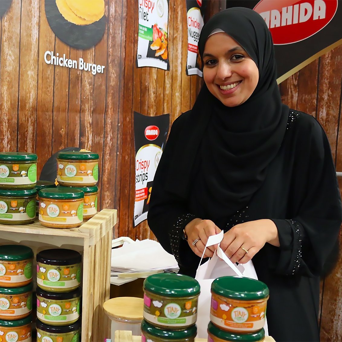 The image features a woman wearing a black hijab, smiling while standing behind a display of jars on a wooden table. The jars are arranged in stacks, showcasing various products, including flavors such as nuggets and chicken burger. The background has a wooden texture, and there are promotional posters or signs visible. The overall setting suggests a market or food exhibition atmosphere.
