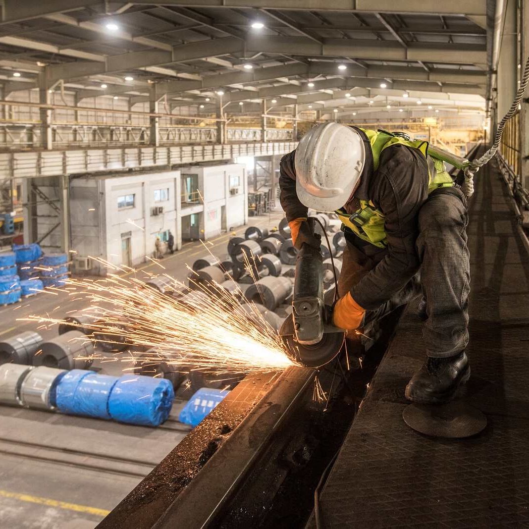 L'image montre un ouvrier en train de travailler dans un environnement industriel, probablement dans une usine. Il utilise une meuleuse, ce qui génère des étincelles. L'ouvrier porte un casque de sécurité et des gants, ce qui indique qu'il suit des mesures de sécurité adéquates. En arrière-plan, on peut voir des bobines de matériaux, ainsi que différents équipements et machines typiques d'un espace de travail industriel. L'éclairage de l'atelier semble naturel, avec des zones bien éclairées et d'autres plus sombres, mettant en valeur le côté dynamique et actif de l'environnement de travail.