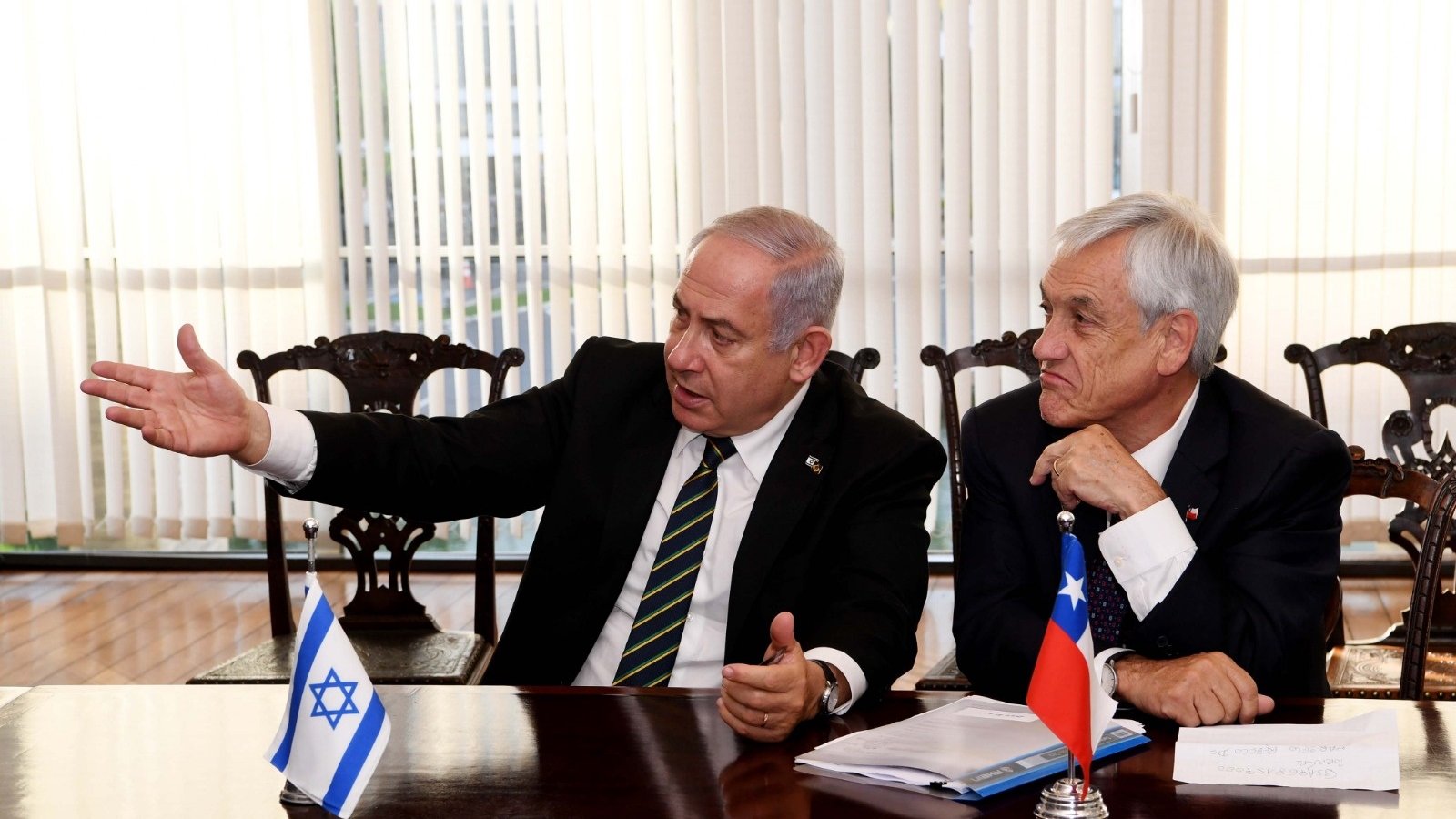 L'image montre deux hommes assis à une table. L'un d'eux, à gauche, gesticule en parlant, tandis que l'autre écoute attentivement. Sur la table se trouvent des documents, et des drapeaux d'Israël et du Chili sont visibles à côté d'eux. L'atmosphère semble sérieuse, suggérant une discussion diplomatique ou politique. Les rideaux derrière donnent une ambiance lumineuse à la scène.