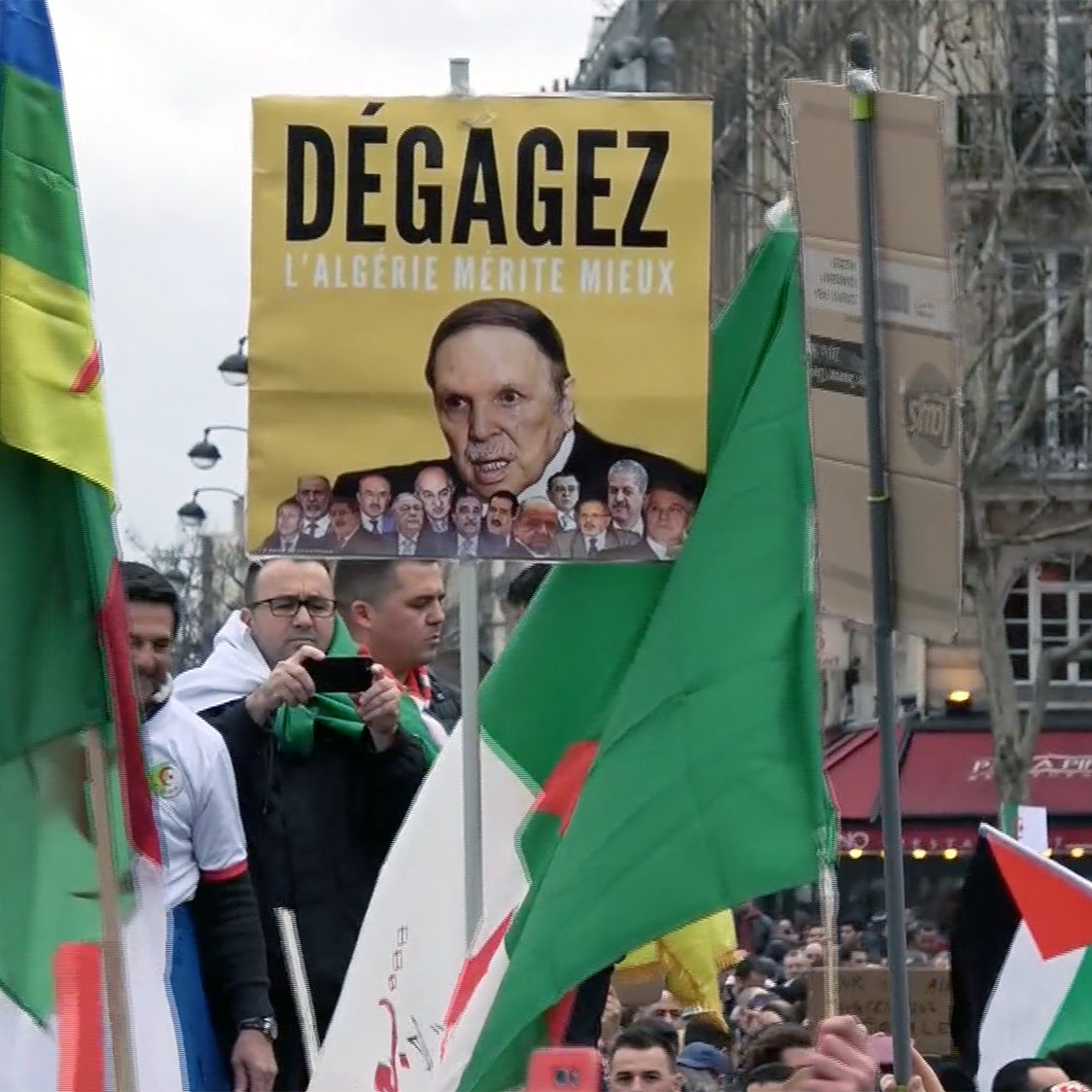 L'image montre une manifestation avec des participants portant des drapeaux. Au centre, un homme tient une grande pancarte sur laquelle est écrit "Dégagez l'Algérie mérite mieux", accompagnée d'une photo d'une personne. Les manifestants semblent exprimer des opinions politiques, et l'atmosphère semble être celle d'une contestation pacifique. Des slogans sur les pancartes reflètent des revendications pour des changements en Algérie.