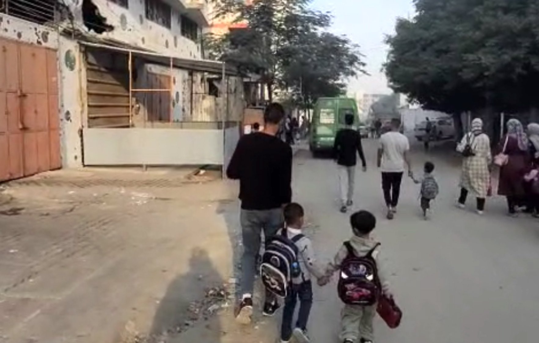 Un uomo cammina con due bambini, mentre altre persone si muovono in una strada cittadina.