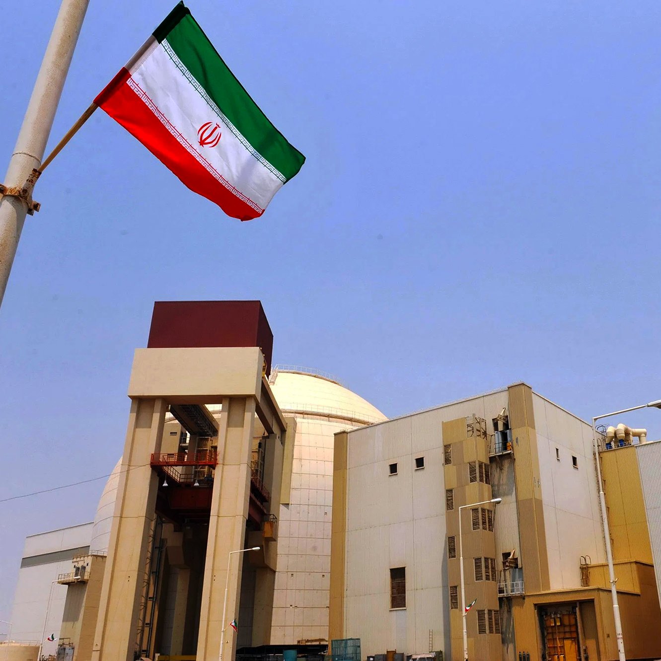 در این تصویر، یک تأسیسات هسته‌ای در ایران دیده می‌شود. در سمت چپ تصویر، پرچم ایران در حال برافراشته شدن است. ساختمان‌ها و برج‌های دودکش در پس‌زمینه قرار دارند و آسمان آبی و شفاف است. تأسیسات به نظر صنعتی و مدرن می‌آیند و نشان‌دهنده فعالیت‌های مرتبط با انرژی هسته‌ای هستند.