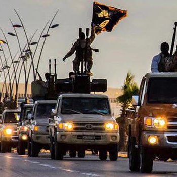 L'image montre une ligne de véhicules tout-terrain circulant sur une route. Certains des véhicules sont équipés d'armes lourdes et portent des drapeaux. Le ciel est partiellement nuageux, ce qui donne une ambiance dramatique à la scène. Des soldats sont visibles à bord des camions, suggérant une présence militaire importante. Des éléments urbains, comme des lampadaires et des bâtiments, sont également présents en arrière-plan.