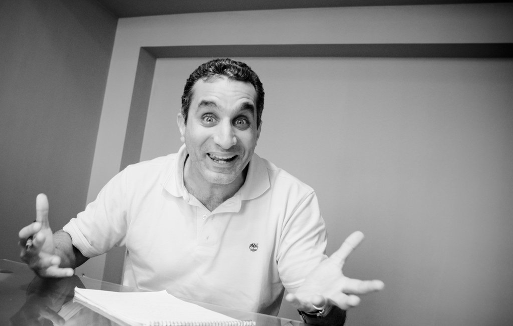 L'image montre un homme assis à une table, avec un air enthousiaste et des gestes expressifs. Il semble parler avec passion, ses bras levés et son visage affichant une grande animation. On peut voir des notes et un petit objet sur la table, suggérant qu'il est en train de discuter ou de partager ses idées. L'image est en noir et blanc, ce qui ajoute une dimension artistique à sa présentation.