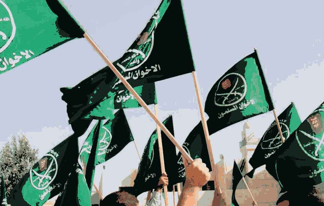 L'image montre un groupe de personnes levant des drapeaux verts. Les drapeaux semblent porter des inscriptions en arabe ainsi que des symboles, indiquant une affiliation à un mouvement ou une organisation spécifique. L'ambiance semble dynamique et engagée, suggérant un rassemblement ou une manifestation. L'arrière-plan indique probablement un espace public ou un lieu symbolique.