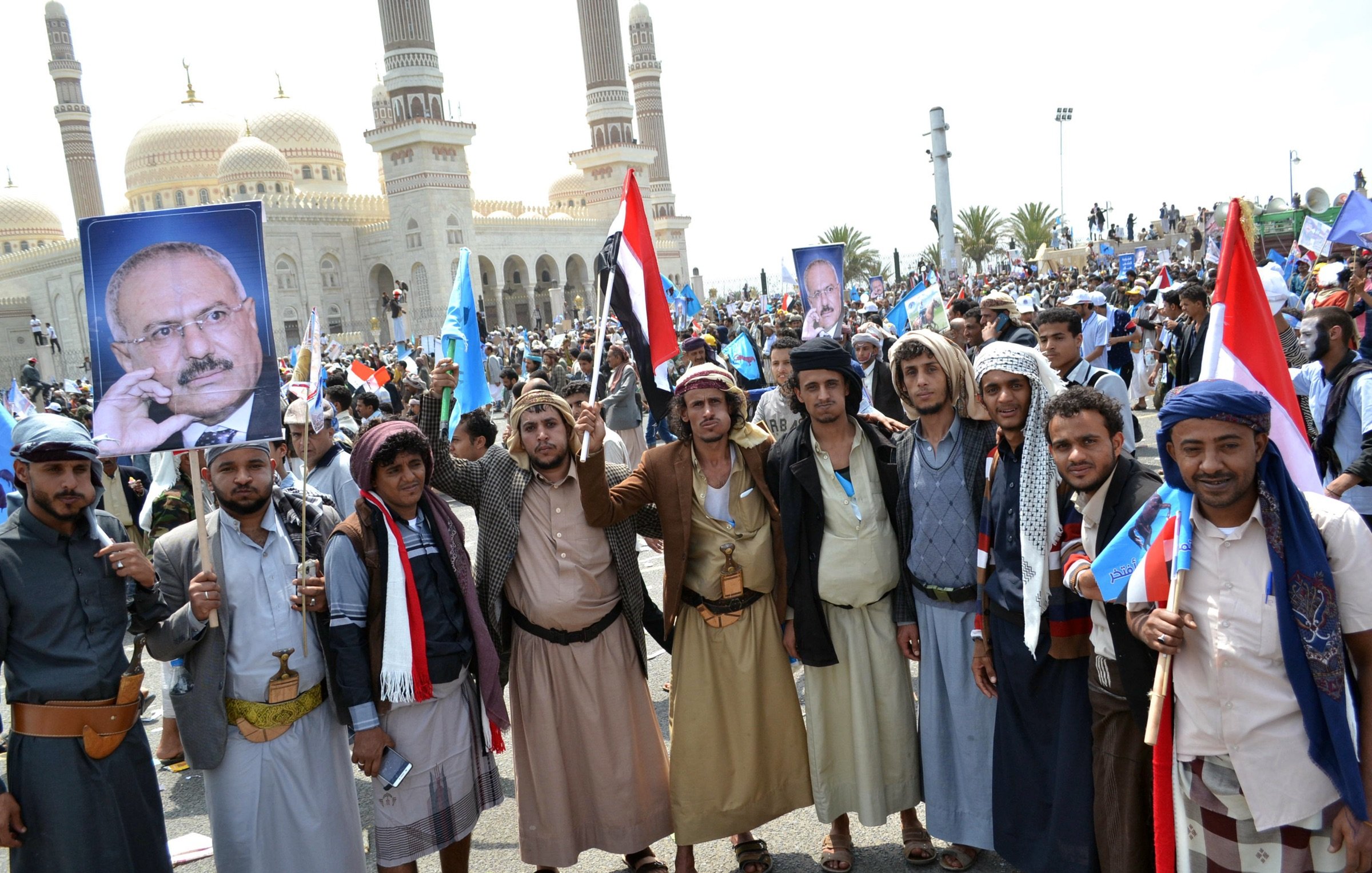 L'image montre un groupe de personnes rassemblées lors d'une manifestation. Elles portent des vêtements traditionnels et tiennent des drapeaux ainsi que des pancartes, y compris une qui affiche un portrait. En arrière-plan, on peut voir une grande mosquée ou un bâtiment avec des dômes, suggérant que l'événement a lieu dans un environnement urbain. L'atmosphère semble festive et engagée, avec une forte mobilisation des participants.