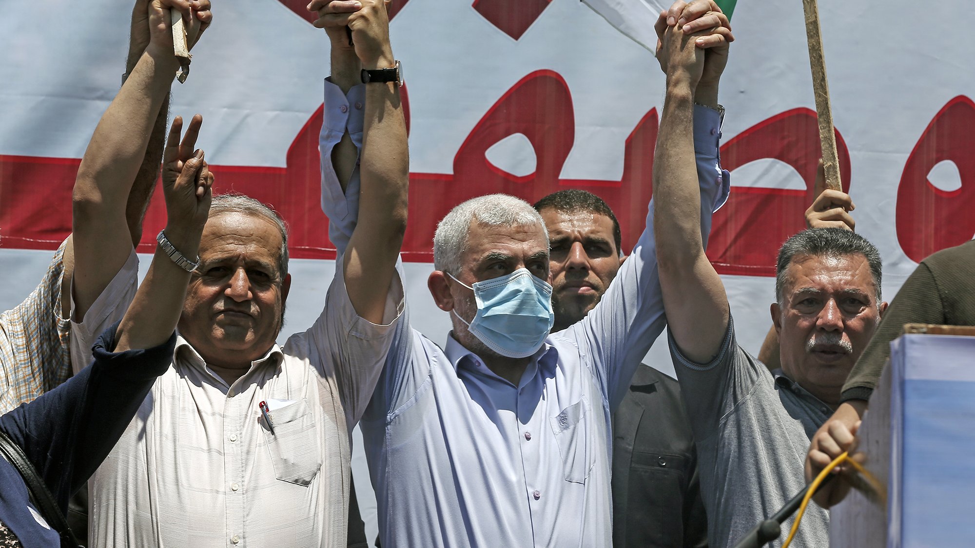 The image depicts a group of men at a public gathering or protest. They are raising their hands in the air, with some holding sticks or flags. The man at the center is wearing a mask and appears to be leading or participating actively in the event. In the background, there is a large banner in Arabic, suggesting a political or social message. The mood of the event seems to be one of solidarity or protest, with participants visibly engaged.