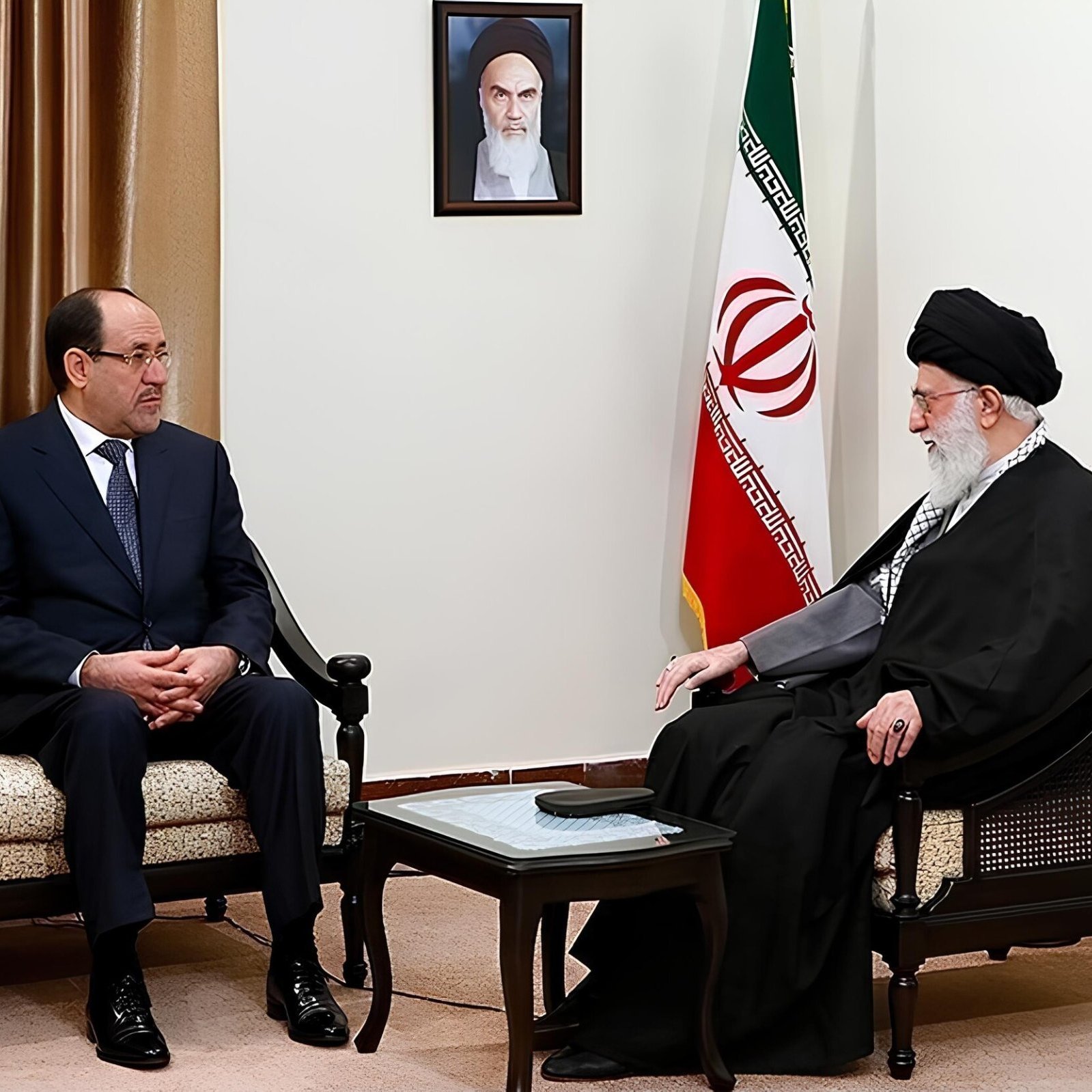 Due uomini seduti in una sala con bandiera iraniana, discutono seriamente.