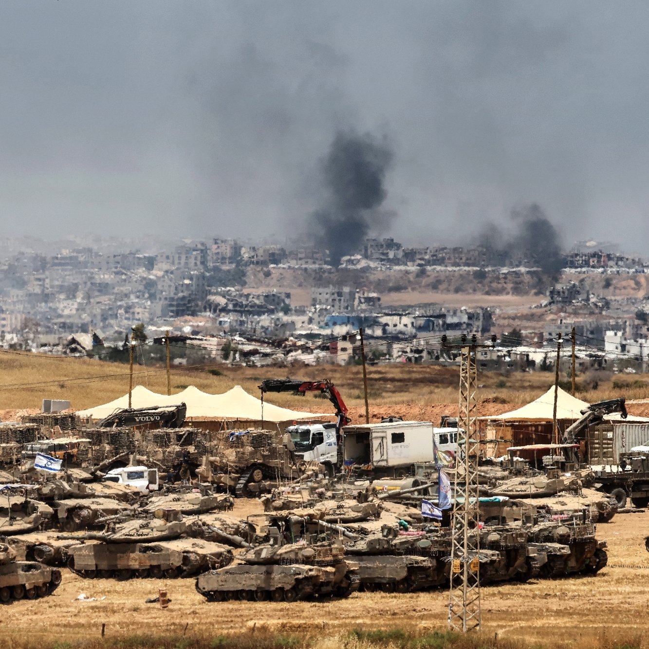 تصویر نشان‌دهنده یک منطقه جنگی با تانک‌ها و دود سیاه از انفجارها در دوردست است.