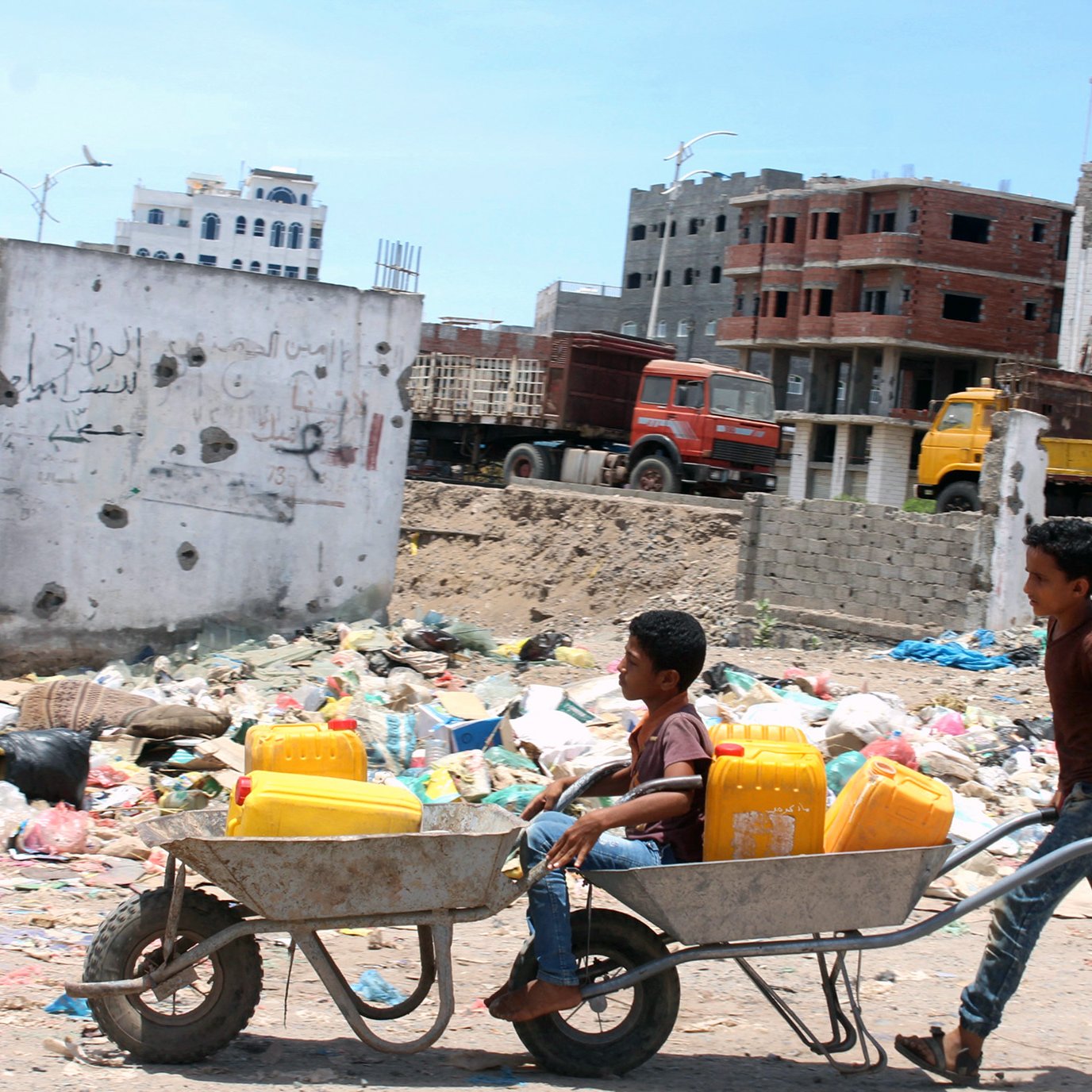 The image shows two boys in a dilapidated urban setting. One boy is pushing a wheelbarrow that carries another boy sitting inside it. The background includes a scene of garbage and rubble, with partially constructed buildings and damaged structures. The environment appears to be neglected, highlighting issues related to poverty and urban decay. The sky is clear, suggesting a bright day.