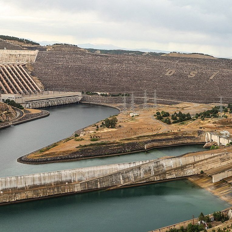 L'image montre une vue panoramique d'un complexe de barrages ou d'une centrale hydroélectrique. On peut voir un grand réservoir d'eau entouré par des collines sur lesquelles se trouvent des structures en béton. Des canaux ou des tunnels d'acheminement de l'eau sont visibles, ainsi que des installations de contrôle des niveaux d'eau. Le paysage est principalement aride, avec quelques zones verdoyantes et une atmosphère dramatique due à un ciel nuageux. Les infrastructures semblent bien entretenues et sont probablement liées à la production d'énergie.