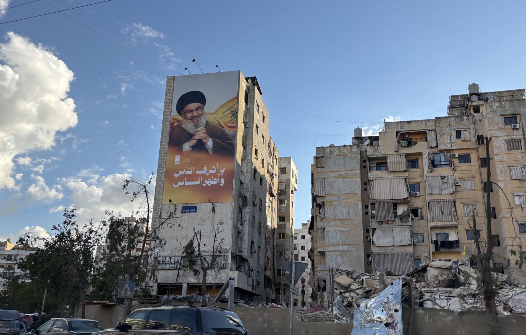 L'image montre un bâtiment en ruines, probablement à la suite d'un conflit ou d'une catastrophe, avec un autre bâtiment voisin encore debout mais en mauvais état. Sur le mur du bâtiment en meilleur état, il y a une grande affiche représentant un homme, probablement un leader ou une figure politique, tenant un symbole de pouvoir. Le ciel est partiellement nuageux, ce qui donne une ambiance contrastée à la scène. Les structures montrent des signes de délabrement, avec des fenêtres manquantes et des balcons en désordre.