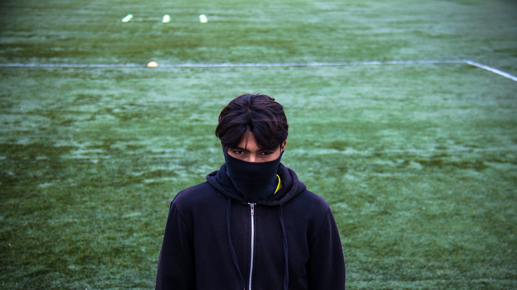 تظهر في الصورة شخصية شاب يقف على أرضية ملعب رياضي مغطاة بالعشب الصناعي. يرتدي الشاب سترة داكنة، ويغطي وجهه بوشاح أو قطعة قماش سوداء، مما يضفي عليه مظهرًا غامضًا. ينظر مباشرة إلى الكاميرا، مما يبرز قوة تعبيره. الخلفية تتكون من اللون الأخضر للأرضية الرياضية، مع بعض العلامات أو الأعلام الموزعة عليها.