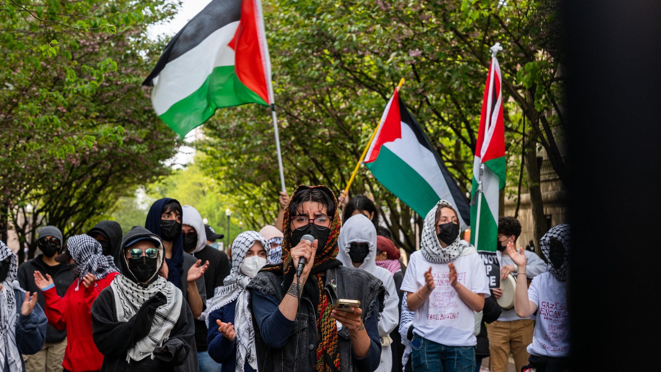 L'image montre une manifestation en plein air où plusieurs personnes sont rassemblées. Au premier plan, une personne prend la parole avec un microphone, vêtue de vêtements sombres et entourée de manifestants portant des masques. Beaucoup des présents arbore des foulards ou keffiehs. En arrière-plan, des drapeaux palestiniens flottent, ajoutant de la couleur à la scène. Les manifestants semblent soutenir une cause, probablement liée à la Palestine, et il y a une atmosphère de solidarité parmi le groupe. Les arbres en fleurs encadrent la scène, créant un contraste avec l'intensité de la manifestation.