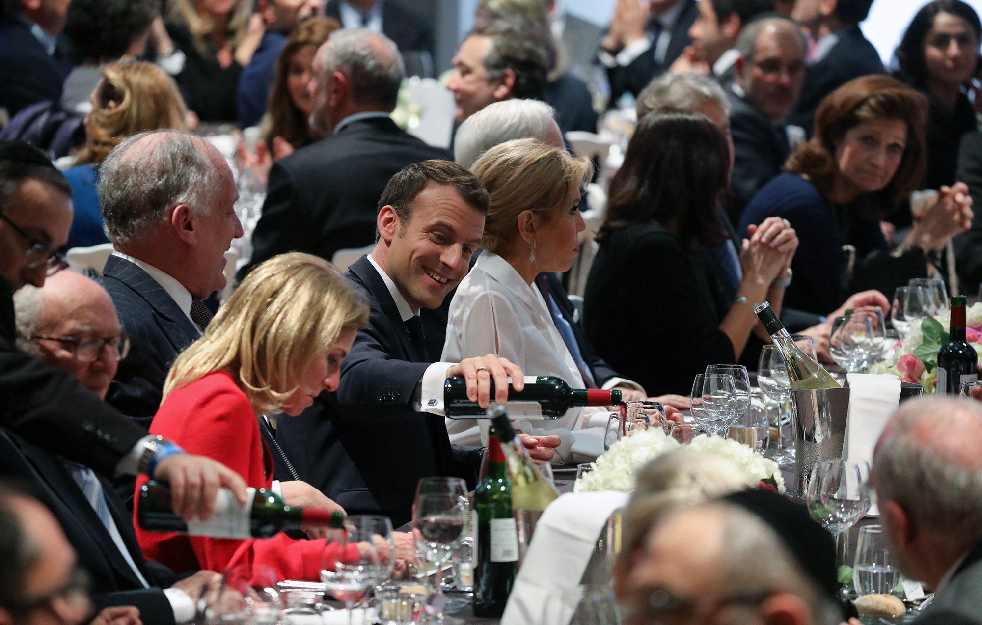 L'image montre une scène de dîner élégant, probablement lors d'un événement officiel. Plusieurs personnes sont assises autour d'une grande table, décorée avec des verres, des bouteilles de vin et des fleurs. Au centre, on aperçoit un homme souriant en train de servir du vin, semblant profiter de l'atmosphère. Les convives sont habillés de manière formelle, et l'ambiance paraît conviviale et animée, avec des conversations visibles parmi les invités.