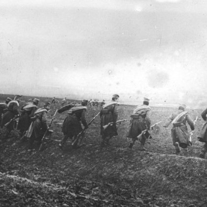 L'image montre un groupe de soldats marchant en formation sur un terrain accidenté. Ils portent des uniformes militaires, avec des chapeaux distinctifs, et semblent se déplacer dans une direction déterminée. Le paysage est marqué par un ciel nuageux et une ambiance sombre, suggérant peut-être un contexte de guerre ou de mouvement stratégique dans un environnement rural. Le sol est boueux, ce qui rend leur avance difficile. L'image est en noir et blanc, renforçant le caractère historique et poignant de la scène.