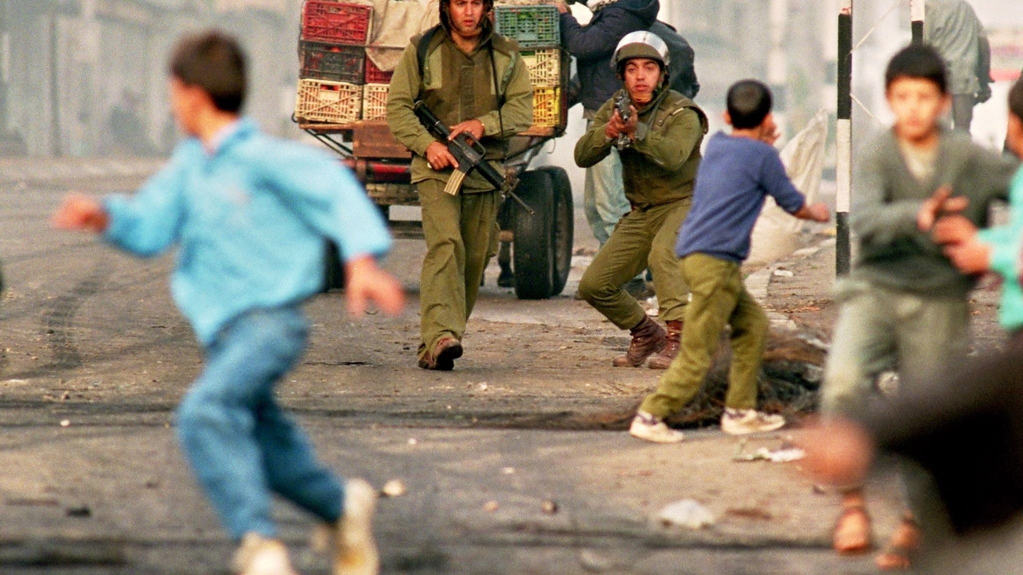L'image montre une scène de tension dans un environnement urbain, où deux soldats sont présents, armés et en position d'action. Un homme, probablement un civil, est également visible, tandis que des enfants et d'autres personnes s'éloignent rapidement de la situation, suggérant un climat de peur ou de chaos. Des véhicules et des baraques sont en arrière-plan, accentuant l'atmosphère de conflit. L'ensemble évoque un contexte de violences ou d'affrontements dans une ville.
