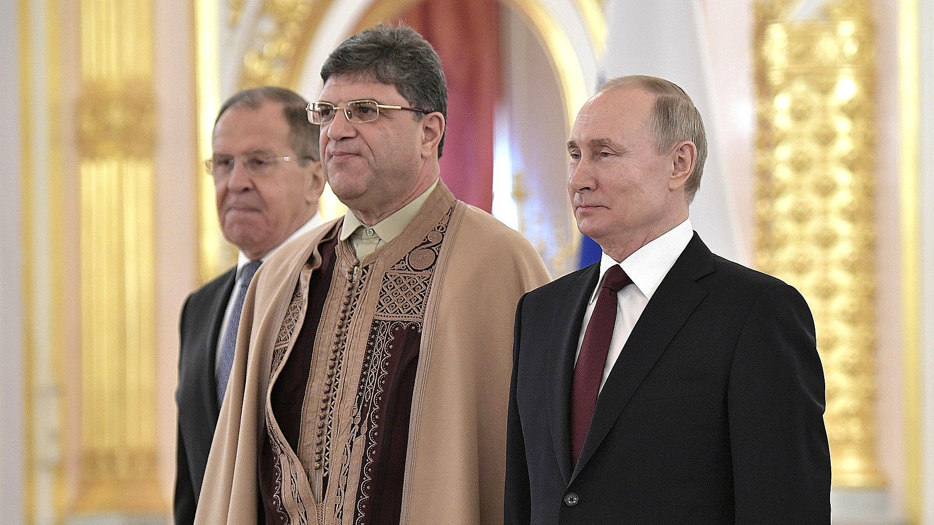 The image shows three men standing together in what appears to be an official setting, likely a ceremonial event. The man on the left is dressed in a suit, while the man in the middle is wearing traditional attire, featuring intricate patterns. The man on the right, who is likely a well-known political figure, is also dressed in a formal suit. The background is ornate, suggesting a formal and significant atmosphere.