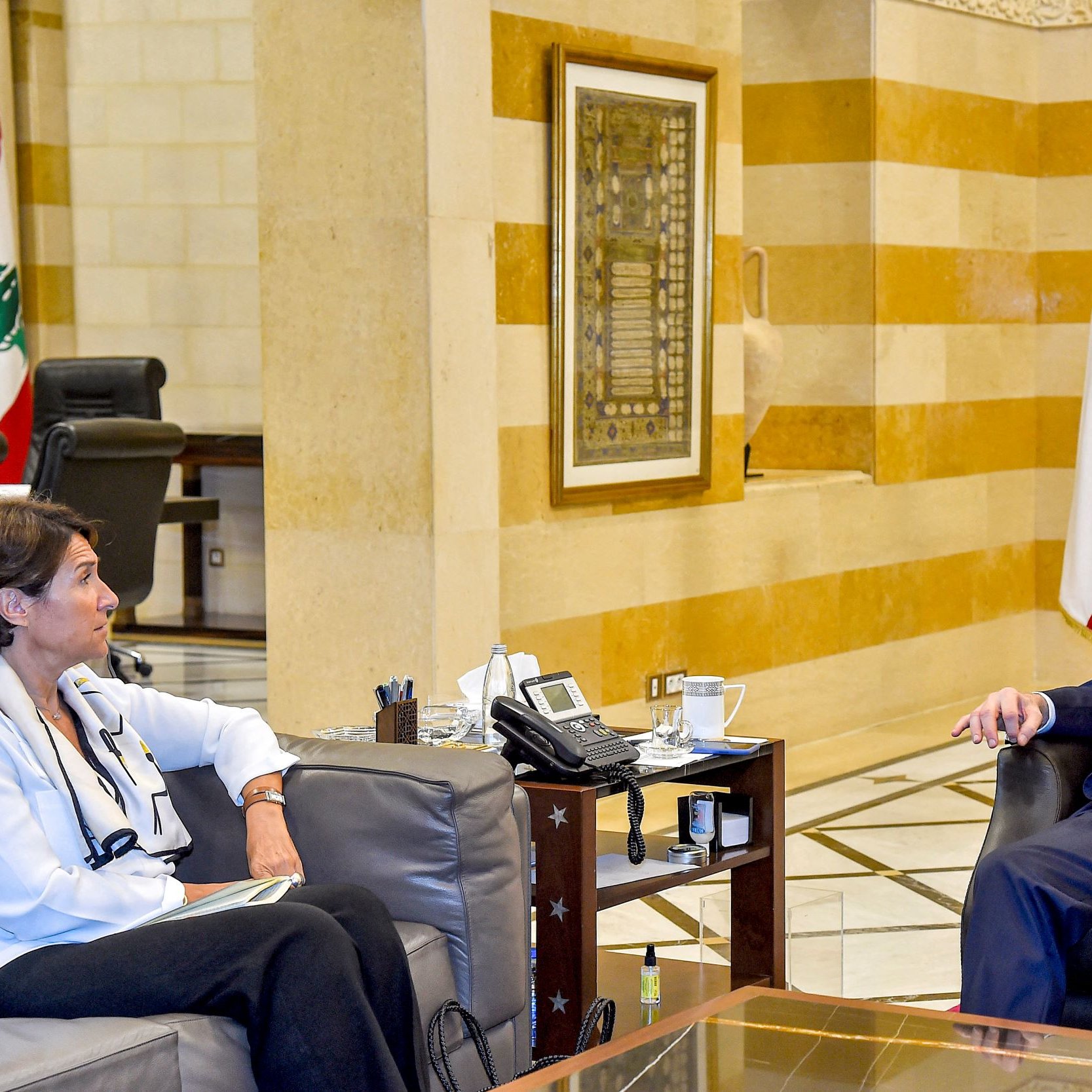 L'image montre une rencontre entre deux personnes assises dans un intérieur soigné, probablement dans un bureau ou une salle de réunion. L'un des individus est un homme en costume, tandis que l'autre est une femme habillée en chemise blanche. Ils semblent engagés dans une discussion, avec des expressions attentives. À l'arrière-plan, on peut apercevoir des drapeaux libanais et une décoration architecturale typique. La scène dégage une ambiance professionnelle et formelle.