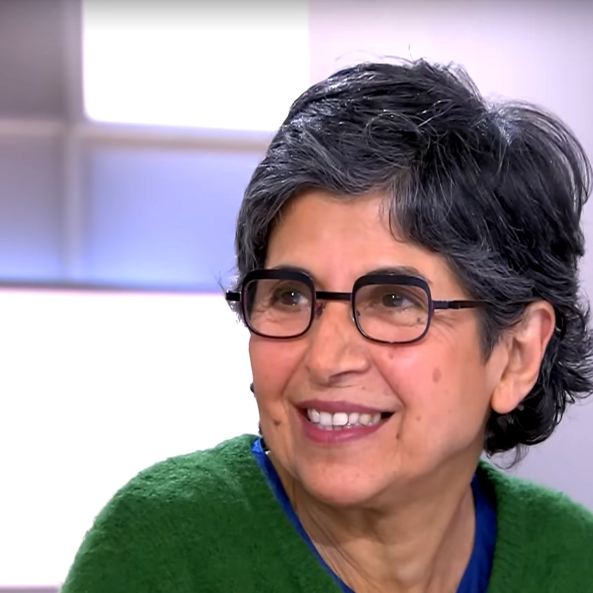 L'image montre une femme souriante avec des cheveux gris et courts. Elle porte des lunettes et un pull de couleur verte. Son expression est amicale et chaleureuse, indiquant qu'elle participe à une conversation. L'arrière-plan est flou, suggérant qu'elle se trouve dans un environnement de studio ou d'interview.