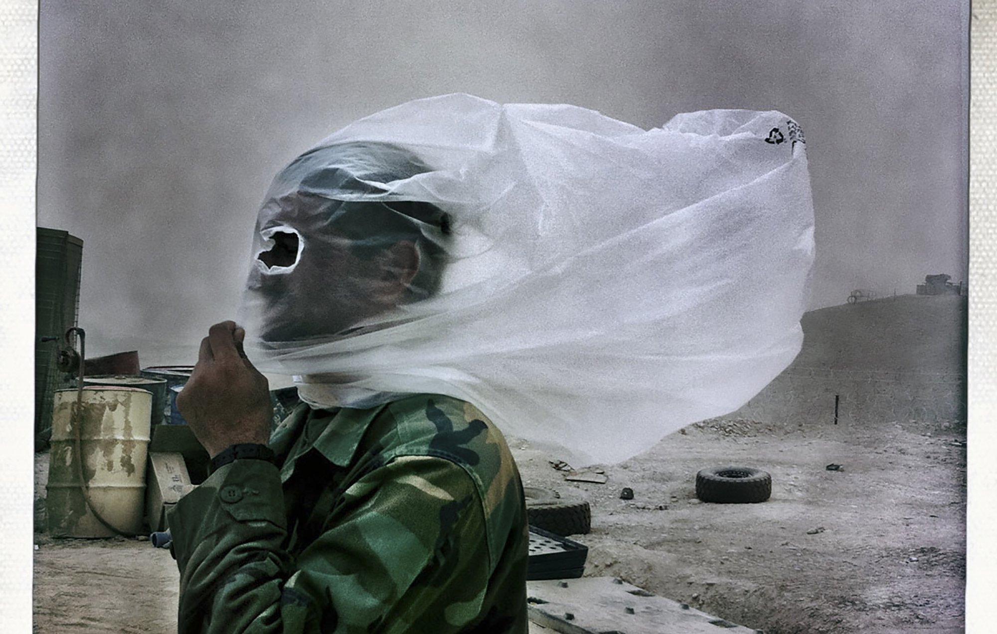 The image depicts a person in military attire, seemingly in a harsh, dusty environment. Their face is partially obscured by a white plastic sheet or bag that is being blown by the wind, creating a dramatic effect. The surroundings appear barren, with hints of debris and tires in the background, suggesting a desolate or rugged terrain. The overall atmosphere conveys a sense of struggle against the elements.
