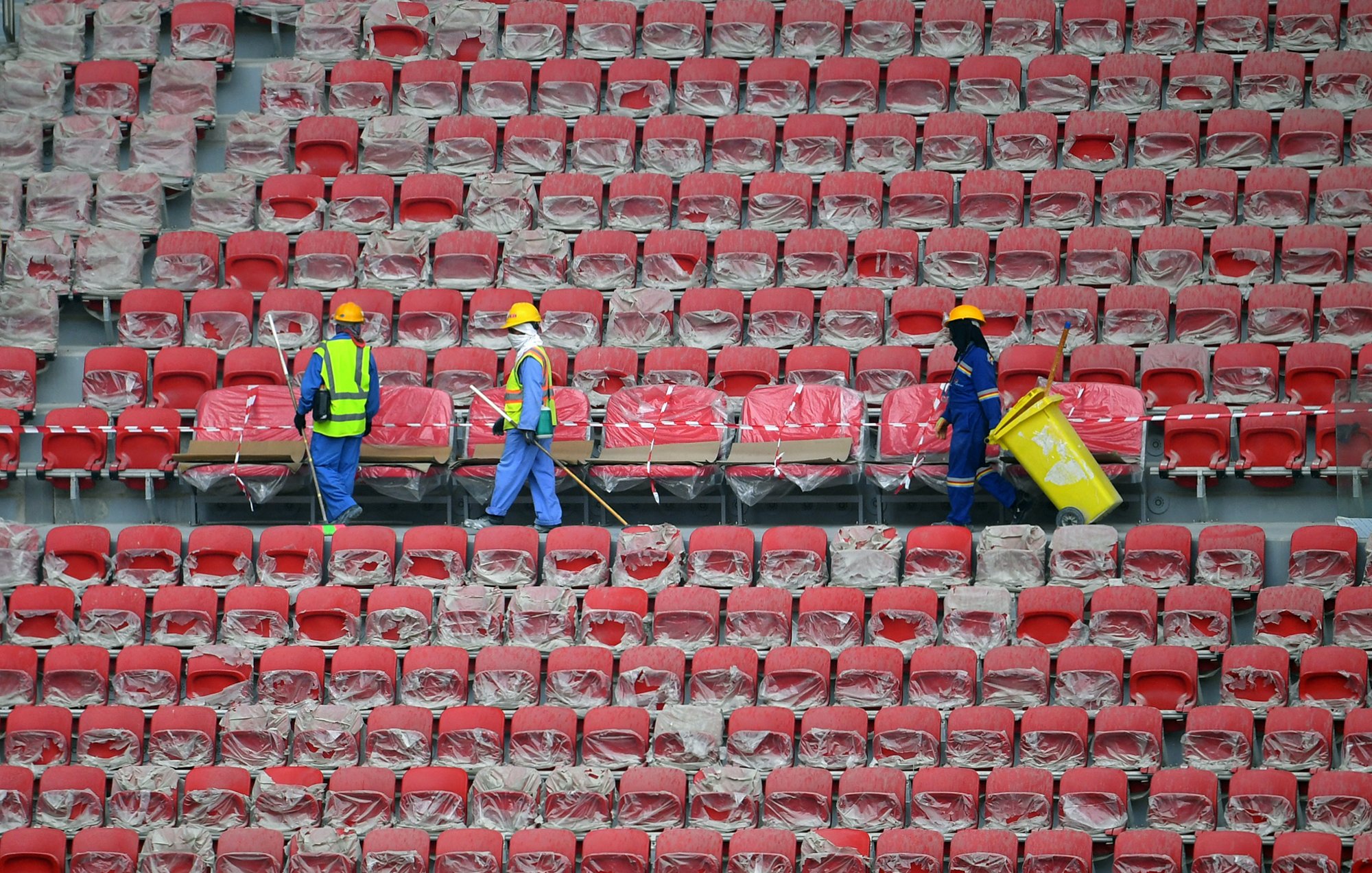 L'image montre des travailleurs dans un stade vide. Ils portent des casques de protection et des gilets réfléchissants. Les sièges du stade sont recouverts de plastique, et les travailleurs semblent être en train de nettoyer ou de préparer les lieux. On peut voir des rangées de sièges rouges. L'atmosphère paraît calme et organisée.