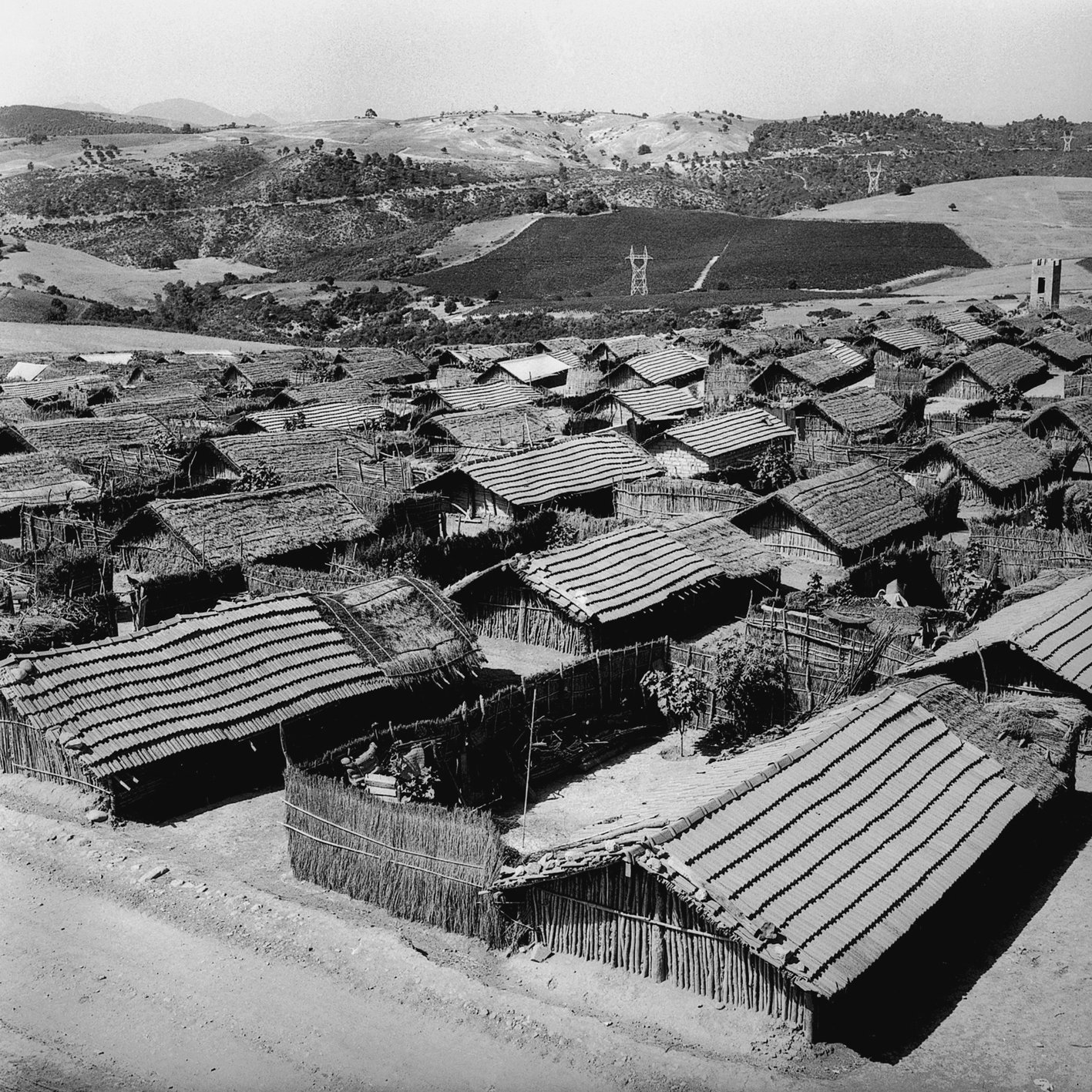 الصورة تُظهر منظرًا عامًّا لقرية صغيرة تتكون من مجموعة من المنازل التقليدية. المنازل تبدو مصنوعة من مواد طبيعية مثل الخشب والقش، مع أسطح مائلة. يتواجد حول القرية تلال خضراء ومناظر طبيعية جميلة، مما يضفي على المشهد طابعًا ريفيًا هادئًا. السماء تبدو صافية، مما يبرز جمال المكان.