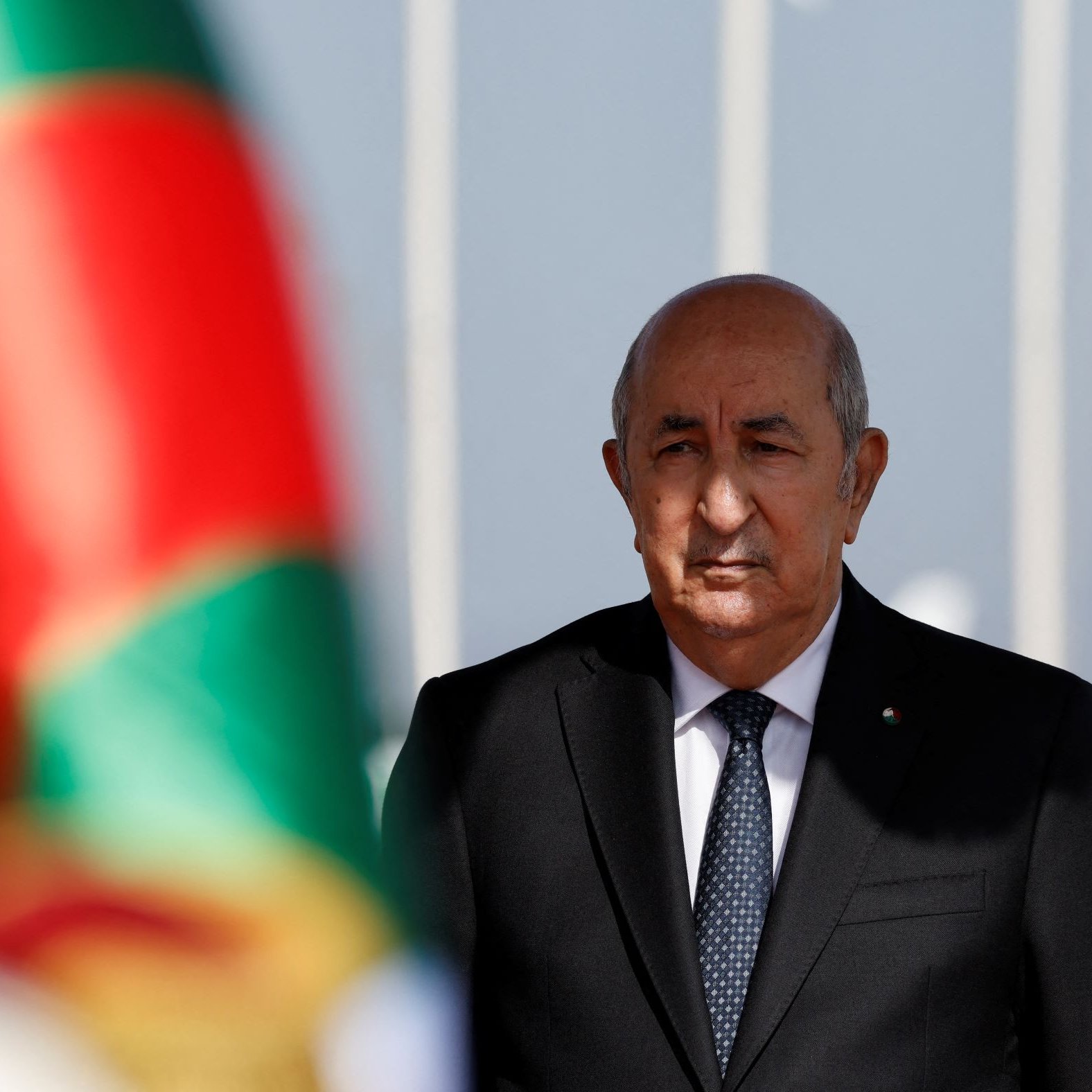 L'image montre un homme en costume sombre, se tenant droit avec une expression sérieuse, en arrière-plan d'un drapeau aux couleurs vives. Le flou devant l'homme suggère qu'il se trouve près d'une cérémonie officielle ou d'un événement important. Le drapeau vert et rouge crée un contraste avec son costume noir.