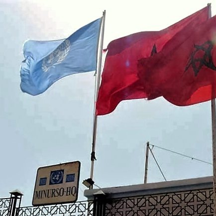 الصورة تظهر علمين مرفوعين على سارية. العلم الأزرق يمثل الأمم المتحدة، بينما العلم الأحمر المزخرف بالنجمة والهلال هو علم المغرب. في الأسفل، يوجد لافتة تشير إلى "مهمة الأمم المتحدة المتكاملة في الصحراء الغربية" (MINURSO)، مما يشير إلى أن الموقع له علاقة بعمليات الأمم المتحدة في هذه المنطقة. السماء في خلفية الصورة تبدو مشمسة.